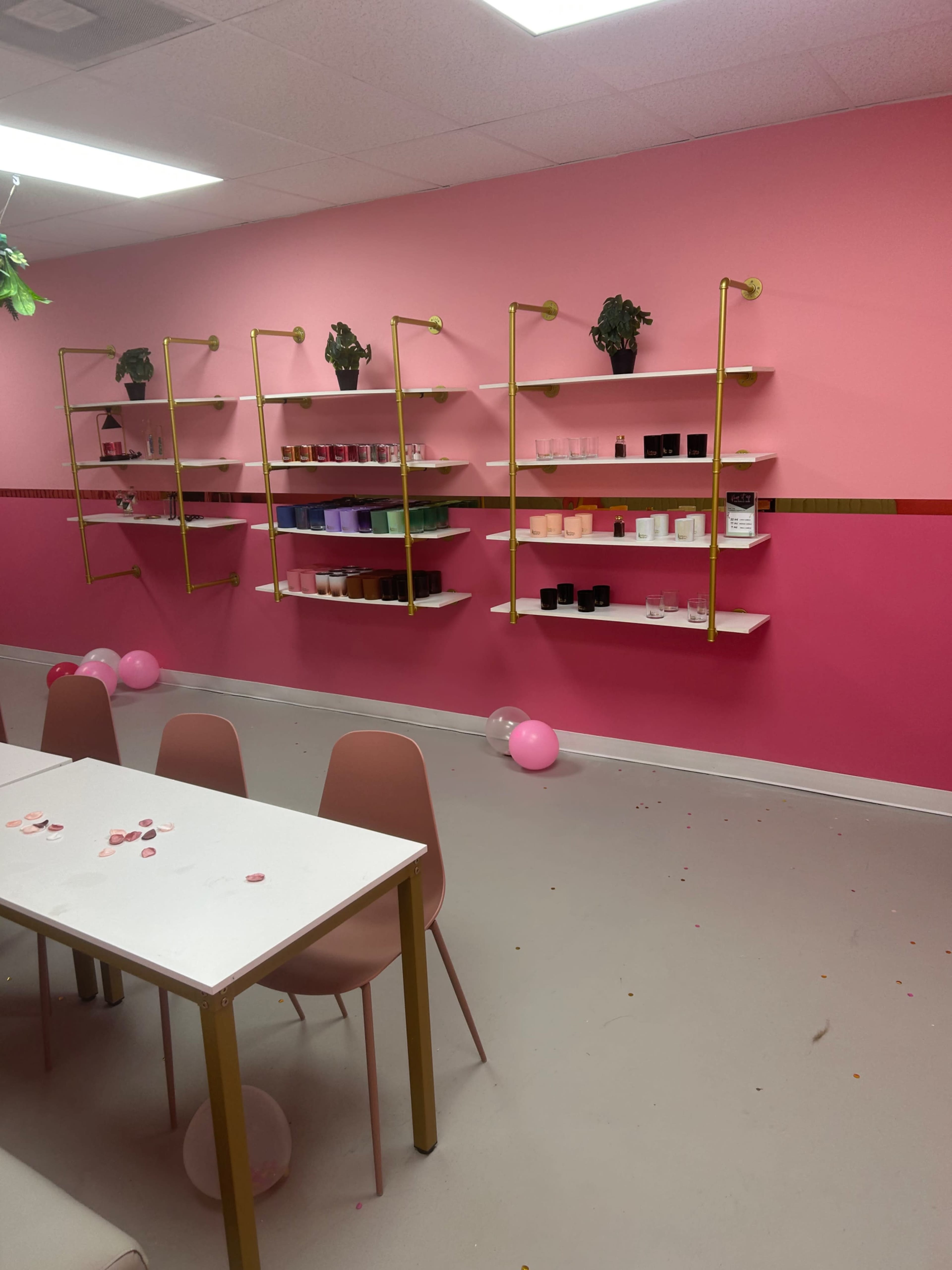 The image shows a brightly colored room with pink walls, featuring white shelves with various items and a table surrounded by chairs, along with scattered decorations and balloons on the floor.