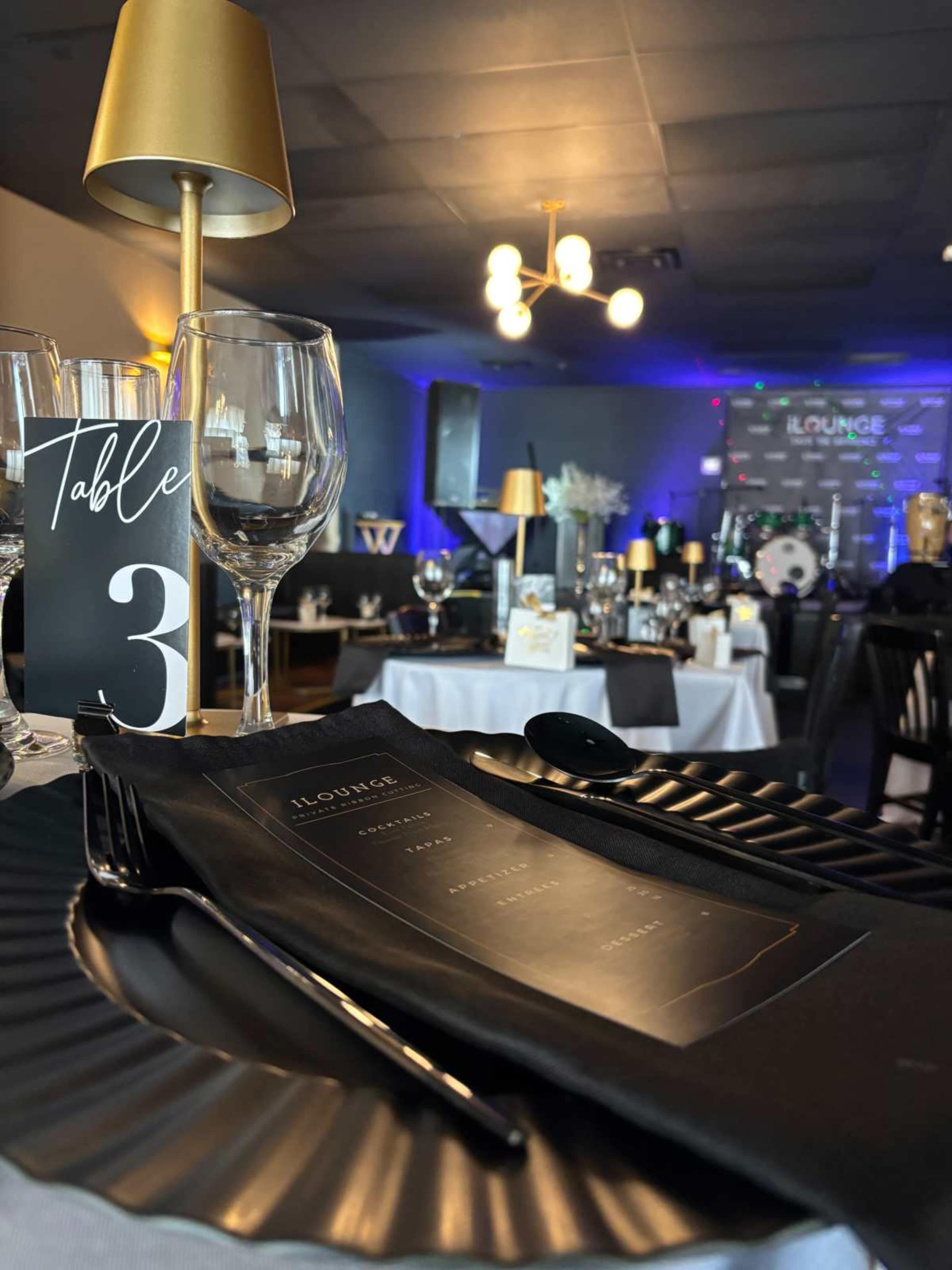 A formal dining setup is displayed on a table with an elegant menu, glassware, and a decorative table number in a softly lit venue featuring a stage in the background.