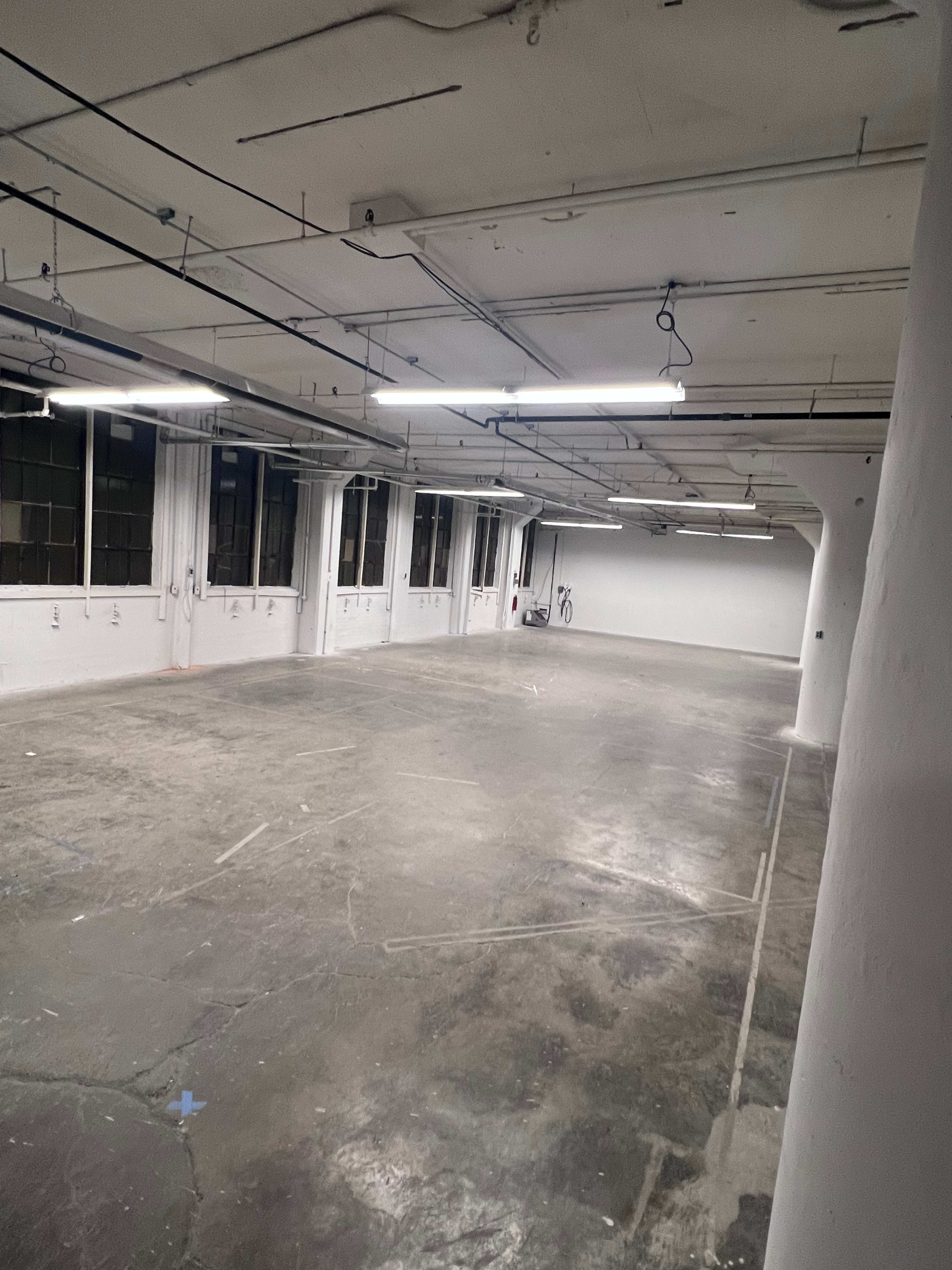 The image shows a large, empty industrial space with concrete floors and exposed ceiling beams, illuminated by fluorescent lights.