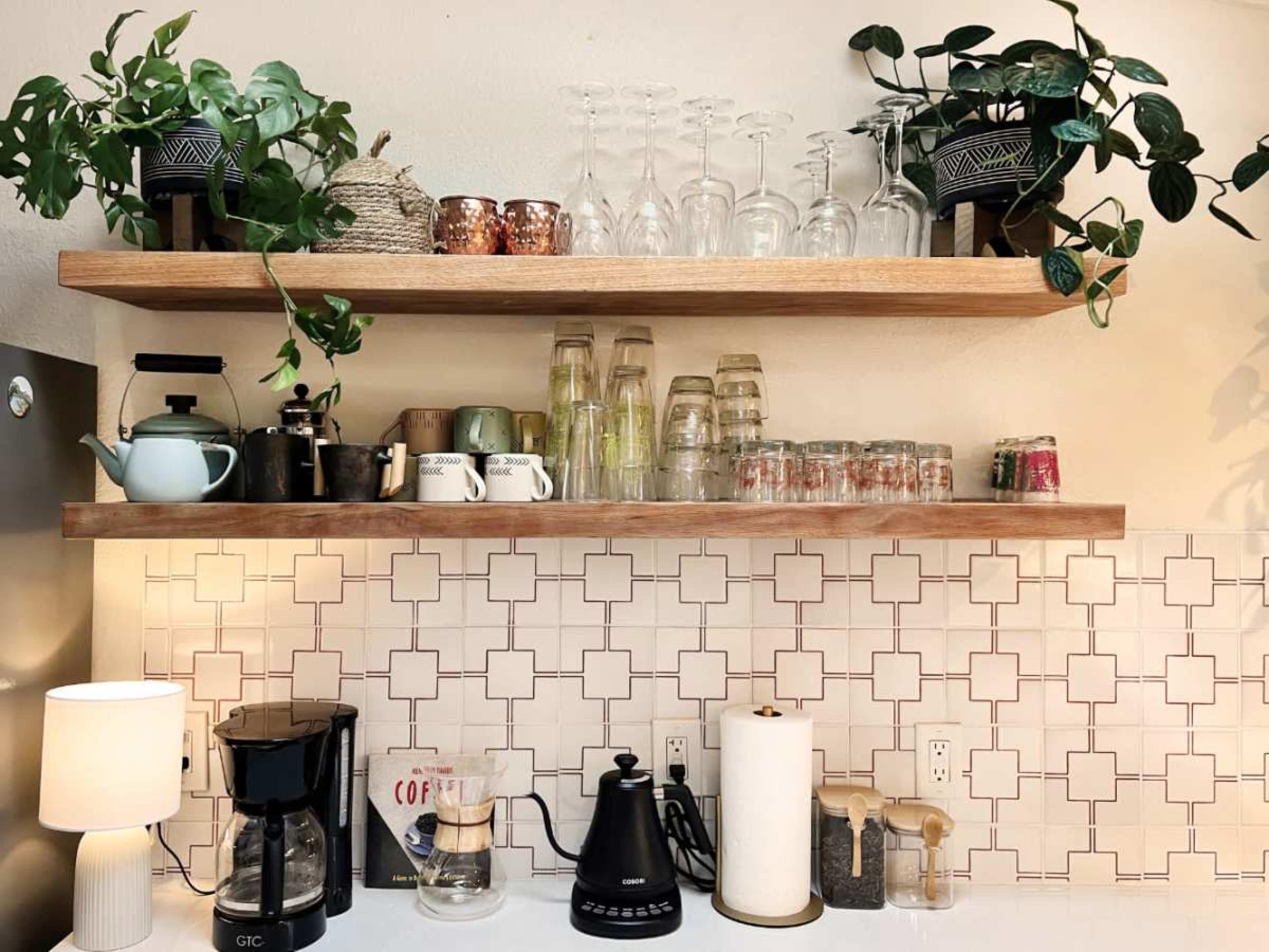 A kitchen counter with wooden shelves displaying glasses, mugs, and plants, alongside a coffee maker and kettle.