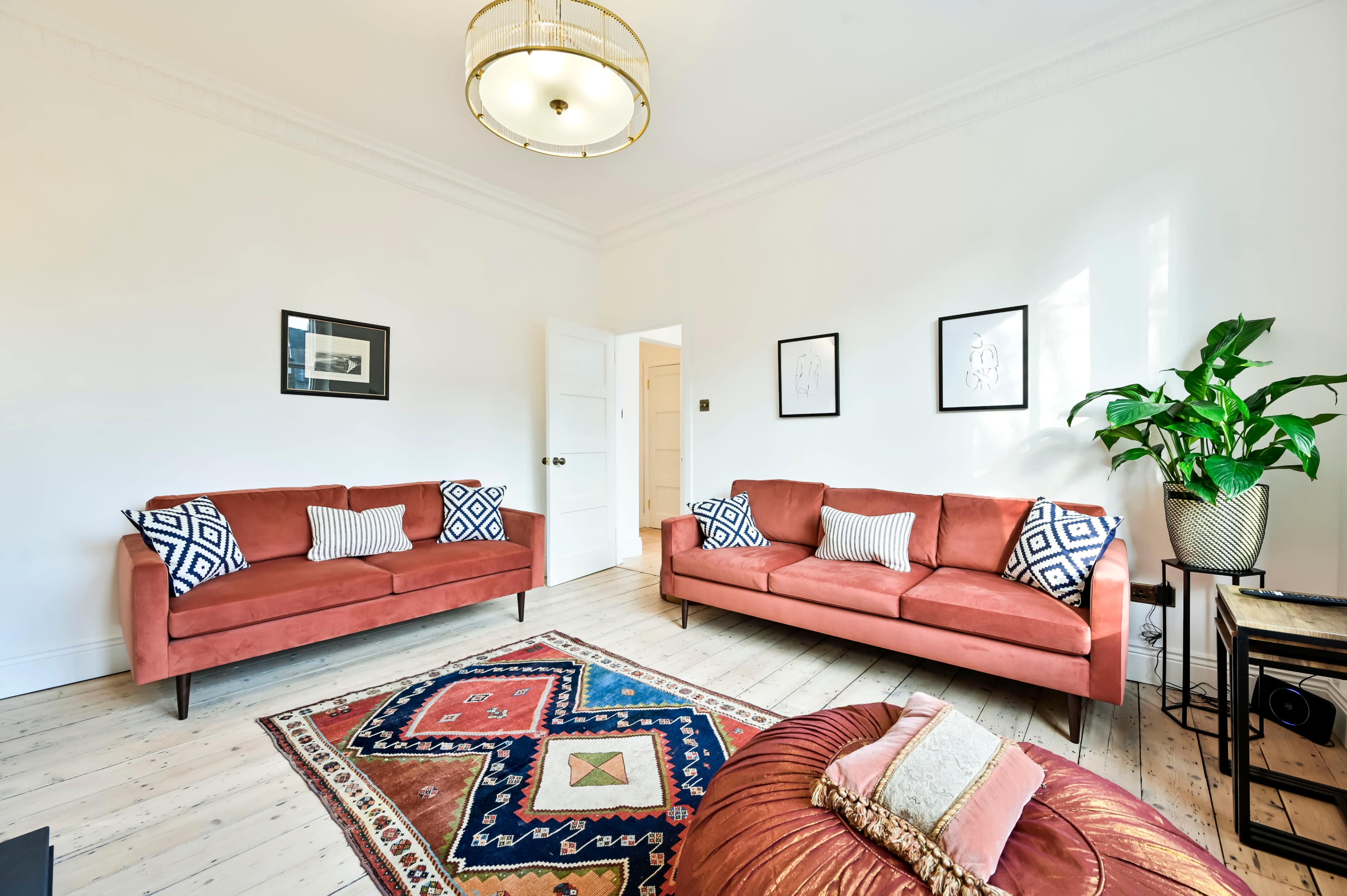 A light-filled living room with two coral-colored sofas facing each other, a patterned rug on the wooden floor, and a large potted plant in the corner.