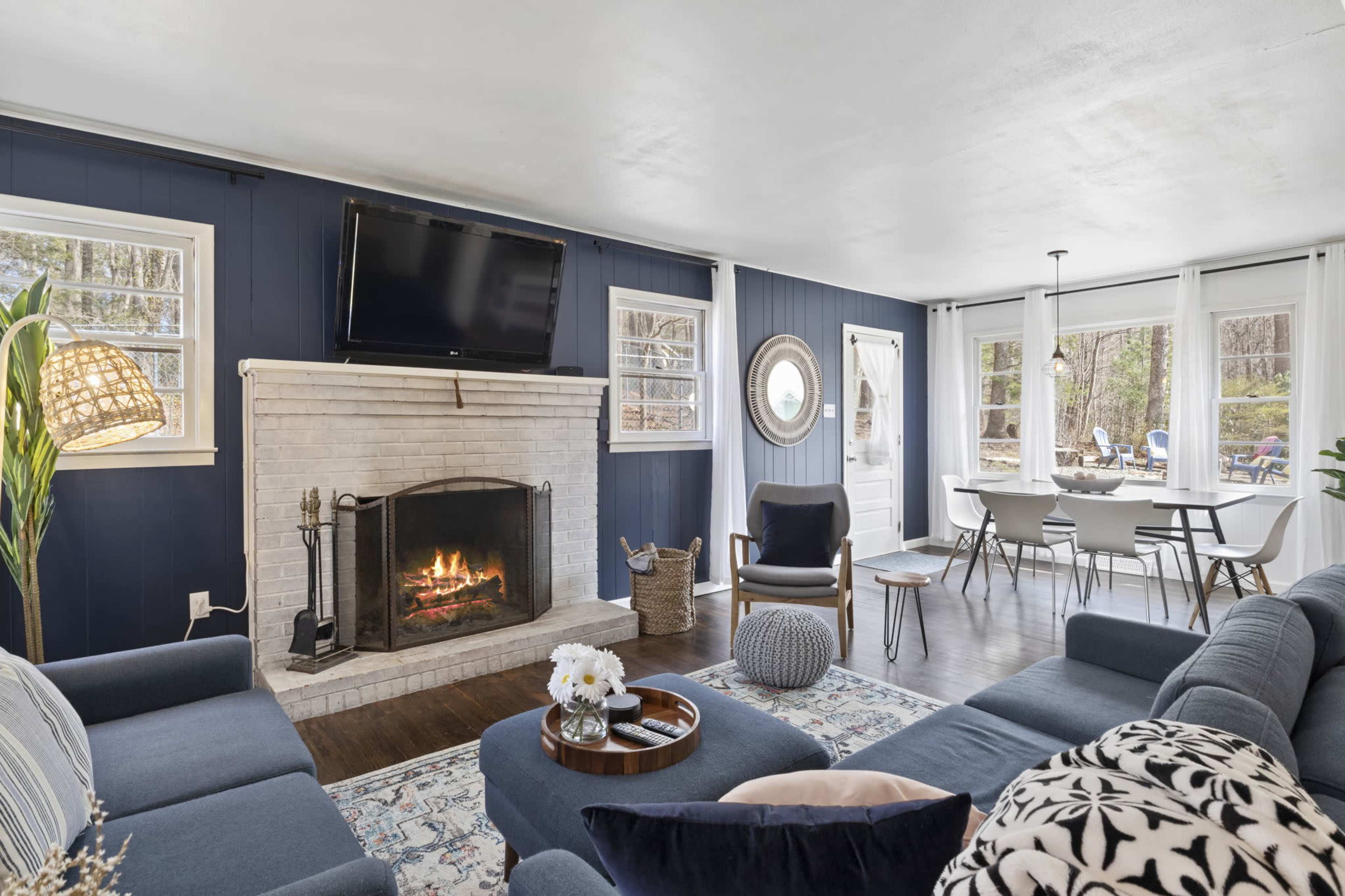 A cozy living room with a fireplace, a mounted TV, and a dining area adjacent to large windows overlooking a natural landscape.