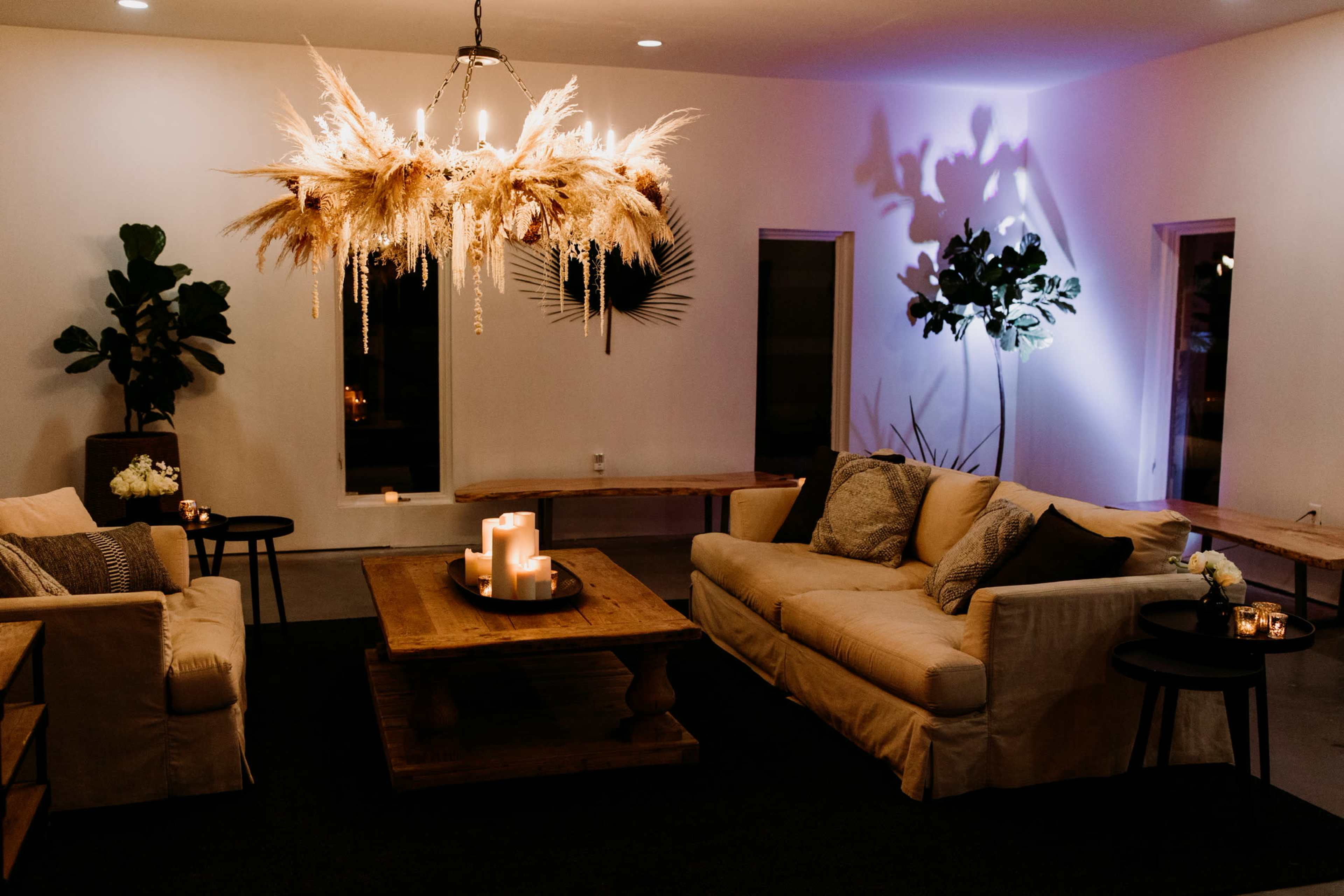 The image depicts a modern living space featuring a large chandelier adorned with dried plants, two sofas facing a wooden coffee table, and ambient lighting creating a cozy atmosphere.