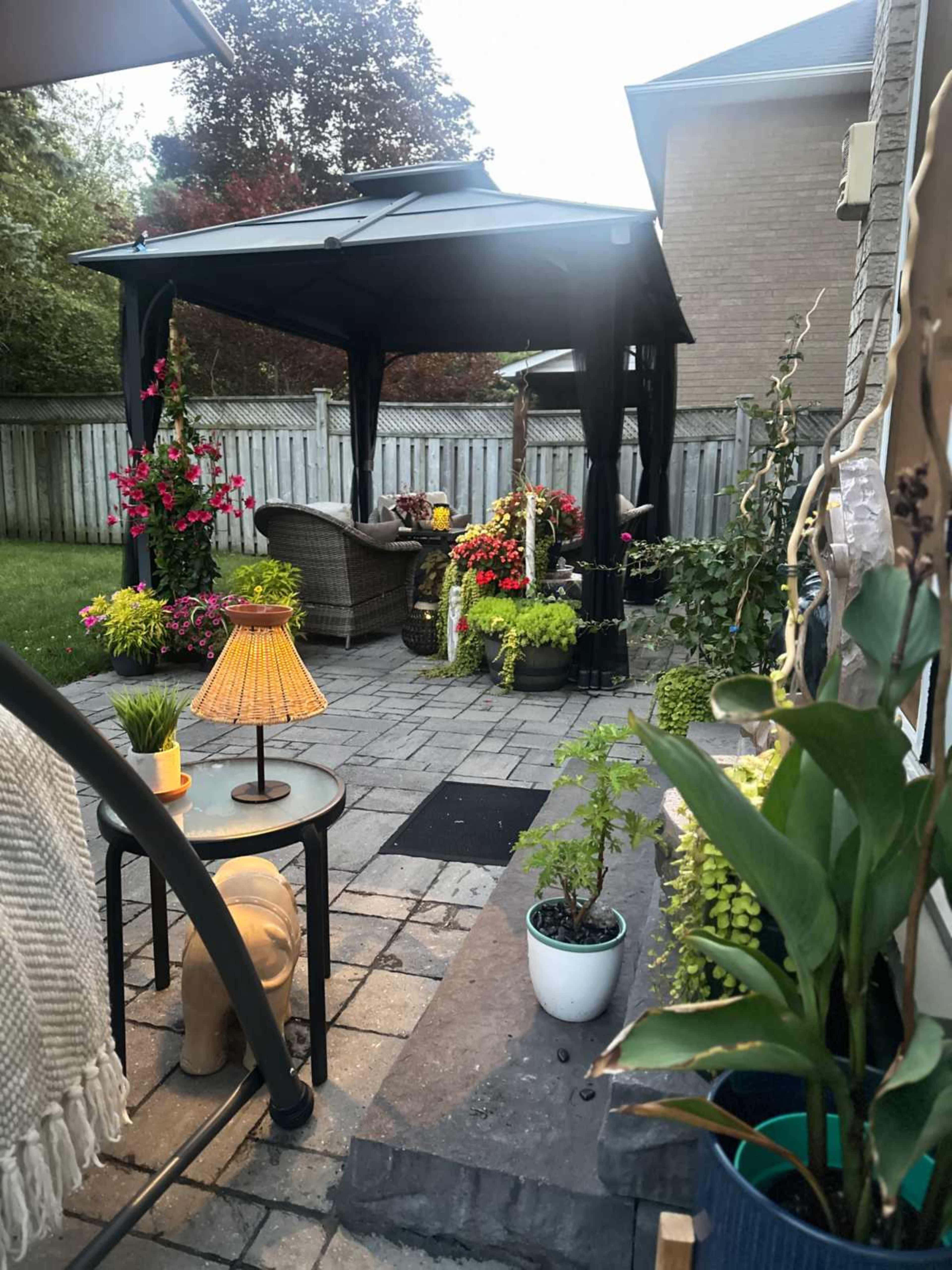 The image shows a cozy backyard patio with a gazebo, surrounded by colorful flowers and plants, and featuring a seating area with furniture and a lamp.