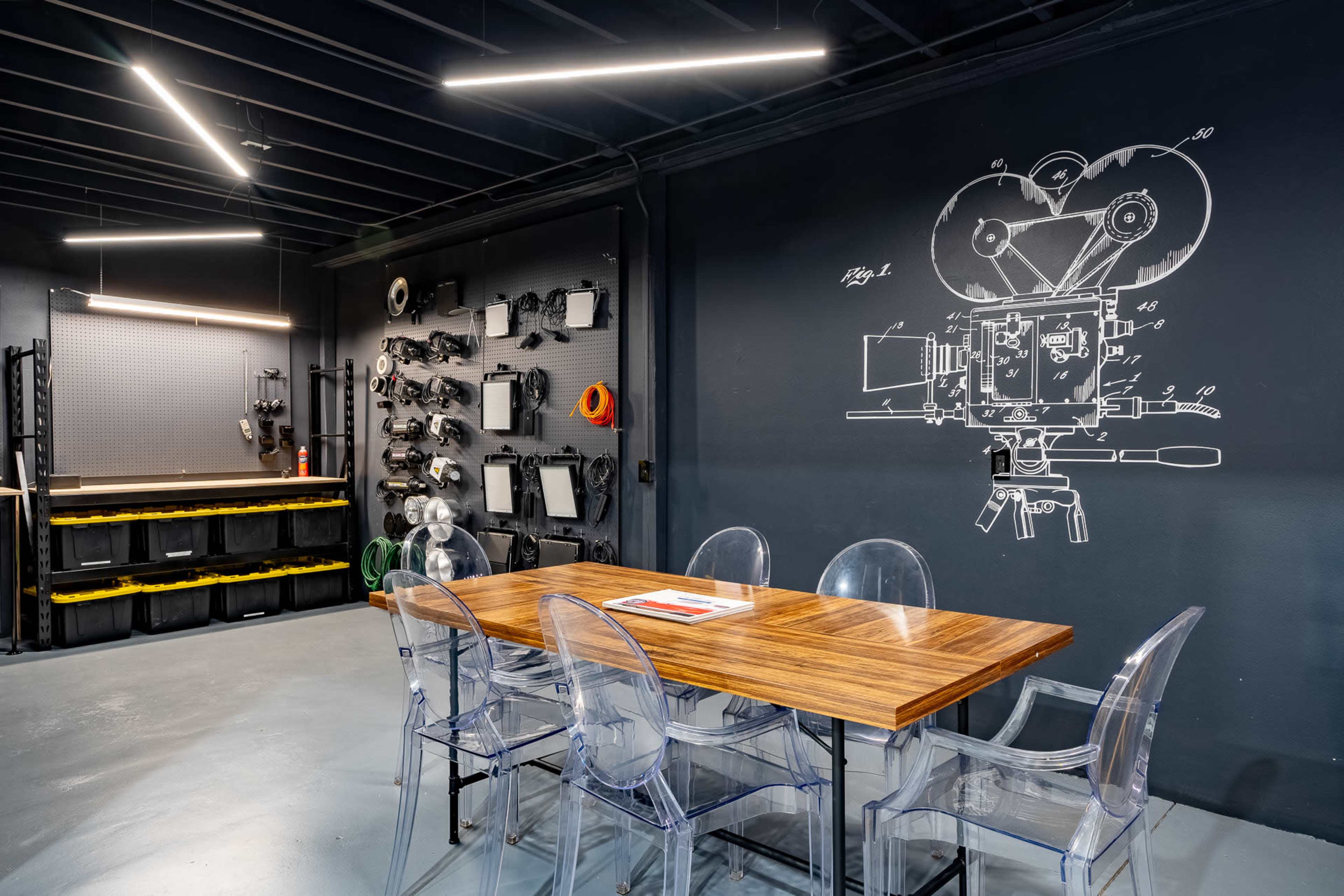 A modern workshop with a long wooden table surrounded by transparent chairs, and walls adorned with tools and a large illustration of a film camera.