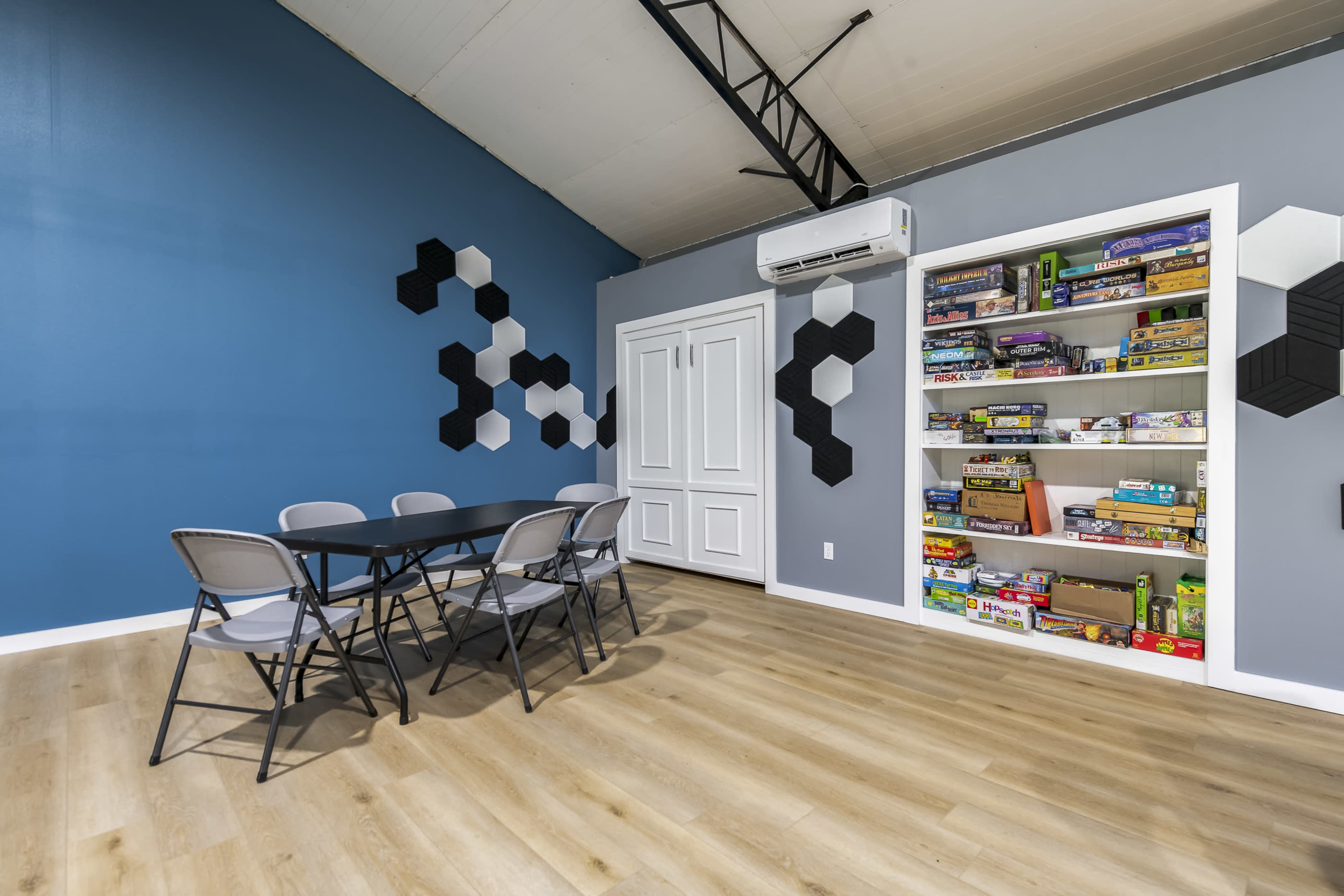 A spacious room features a table with six chairs, hexagonal wall decorations, and a bookshelf filled with board games.