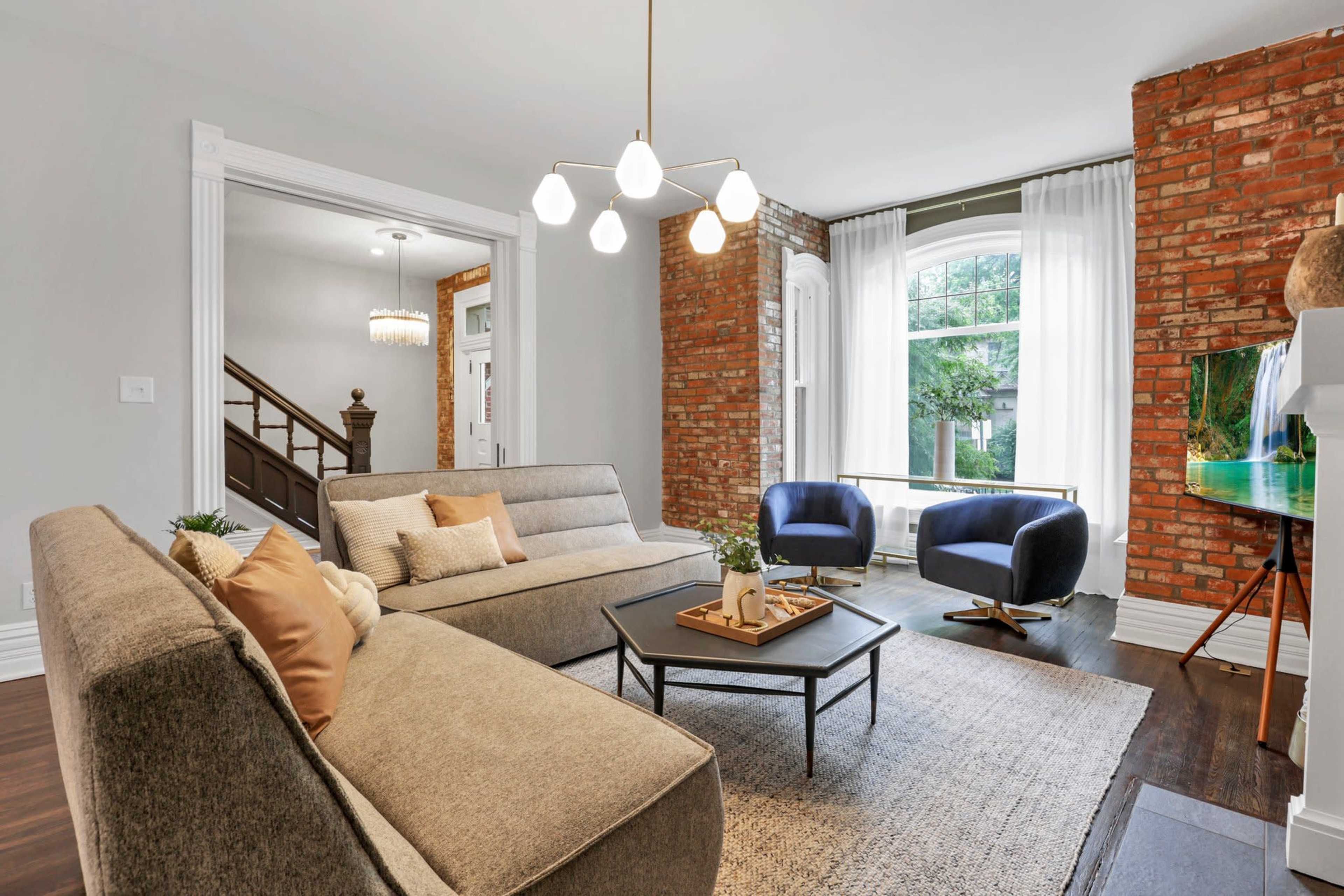 The living room features a modern sectional sofa, two armchairs, a coffee table, and exposed brick walls with a large window providing natural light.