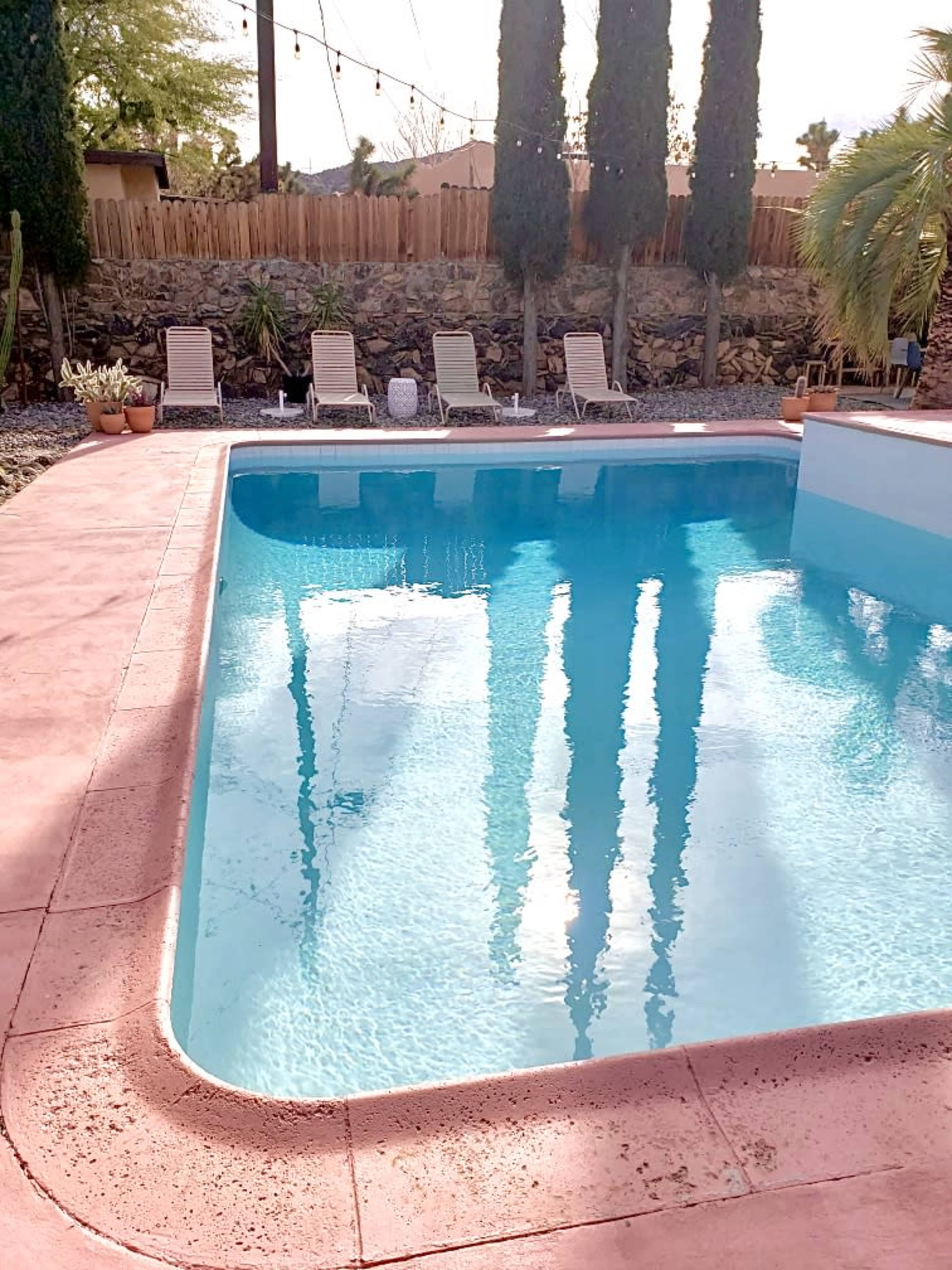 A swimming pool with clear water reflects the surrounding trees and lounge chairs in a landscaped outdoor setting.