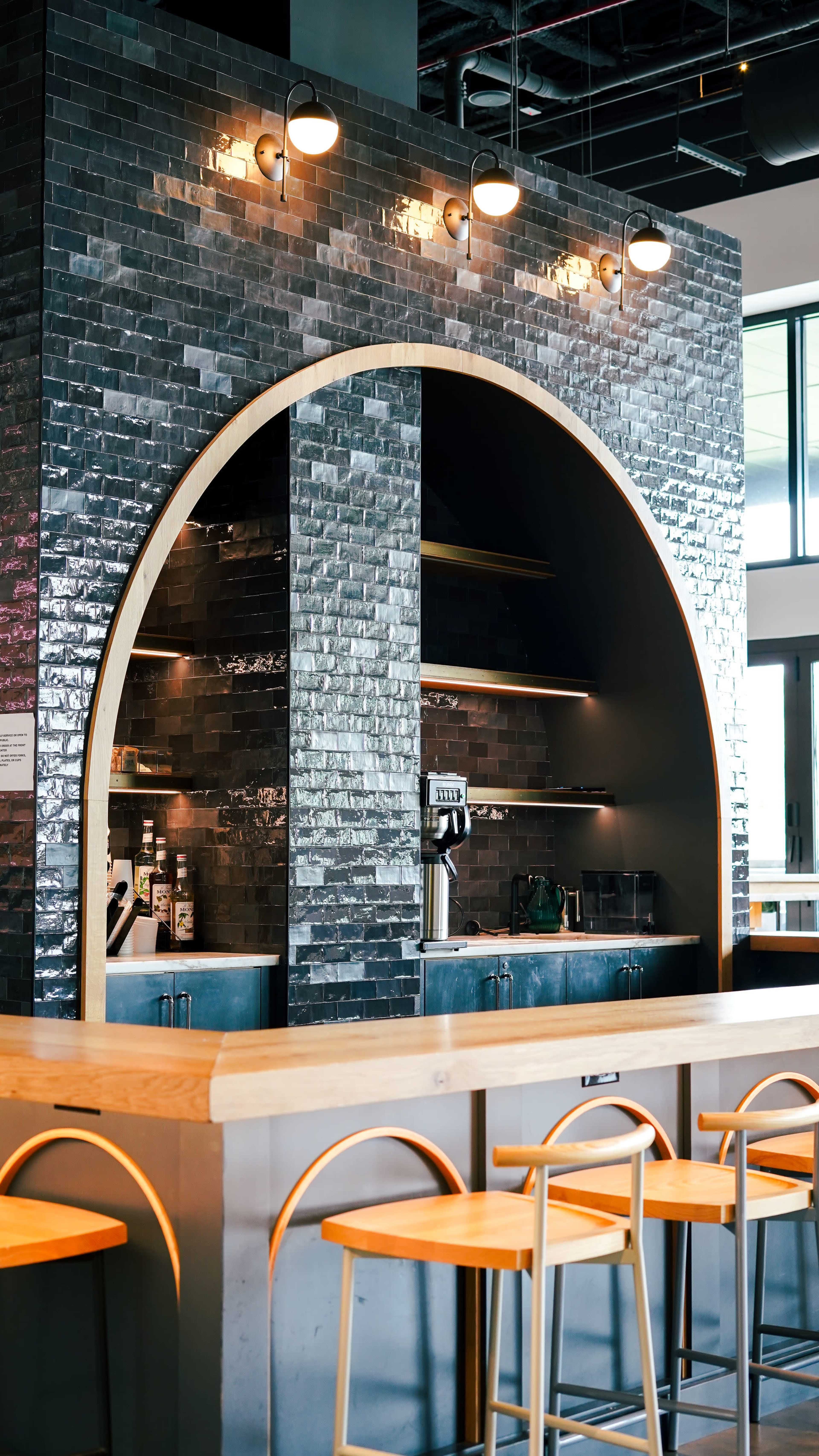 The image shows a modern coffee shop with an arched brick wall featuring shelves and a counter with wooden seating.