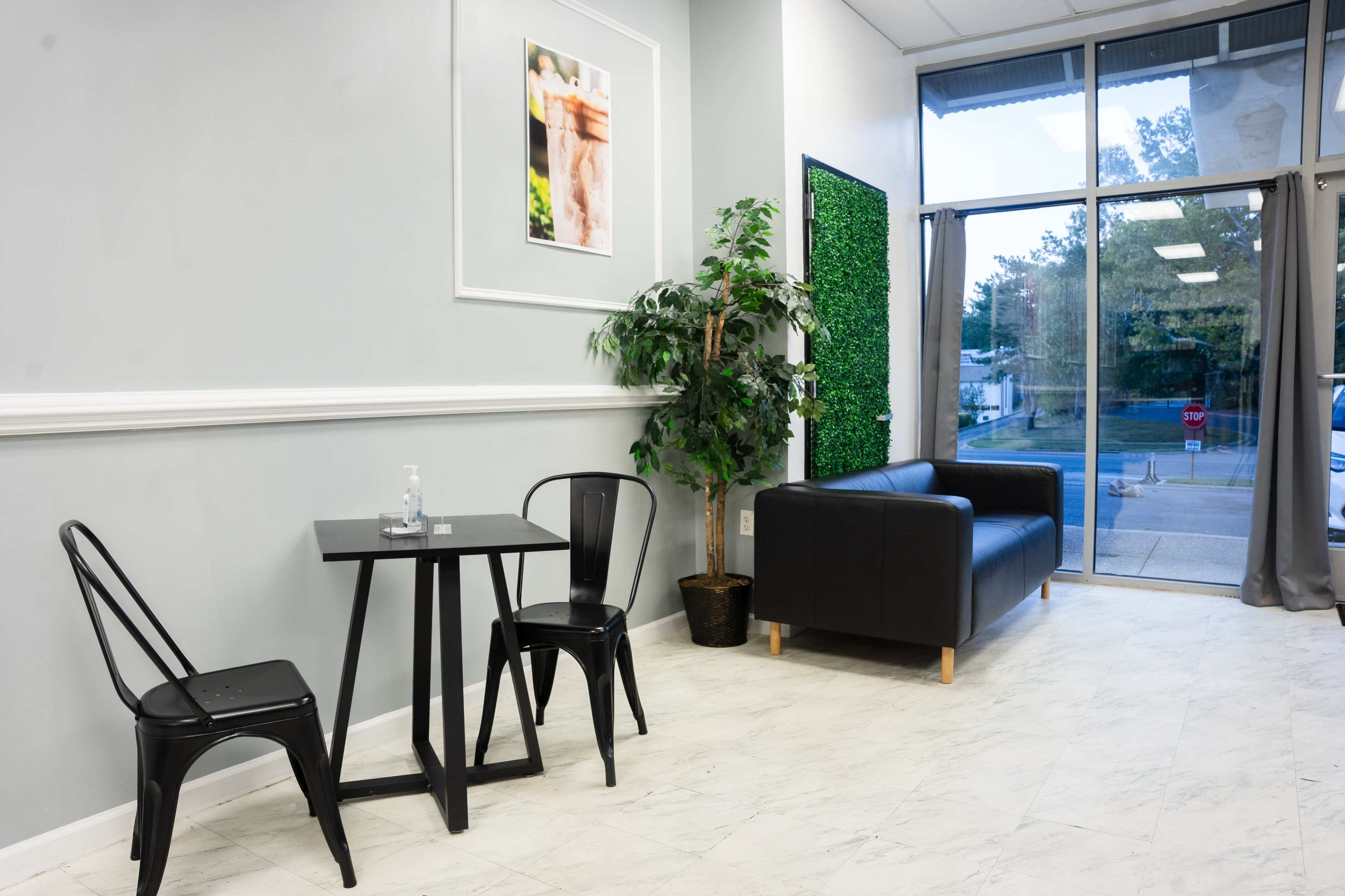 The image shows a modern interior of a waiting area featuring a black sofa, a small table with two chairs, and a decorative plant against a light gray wall.
