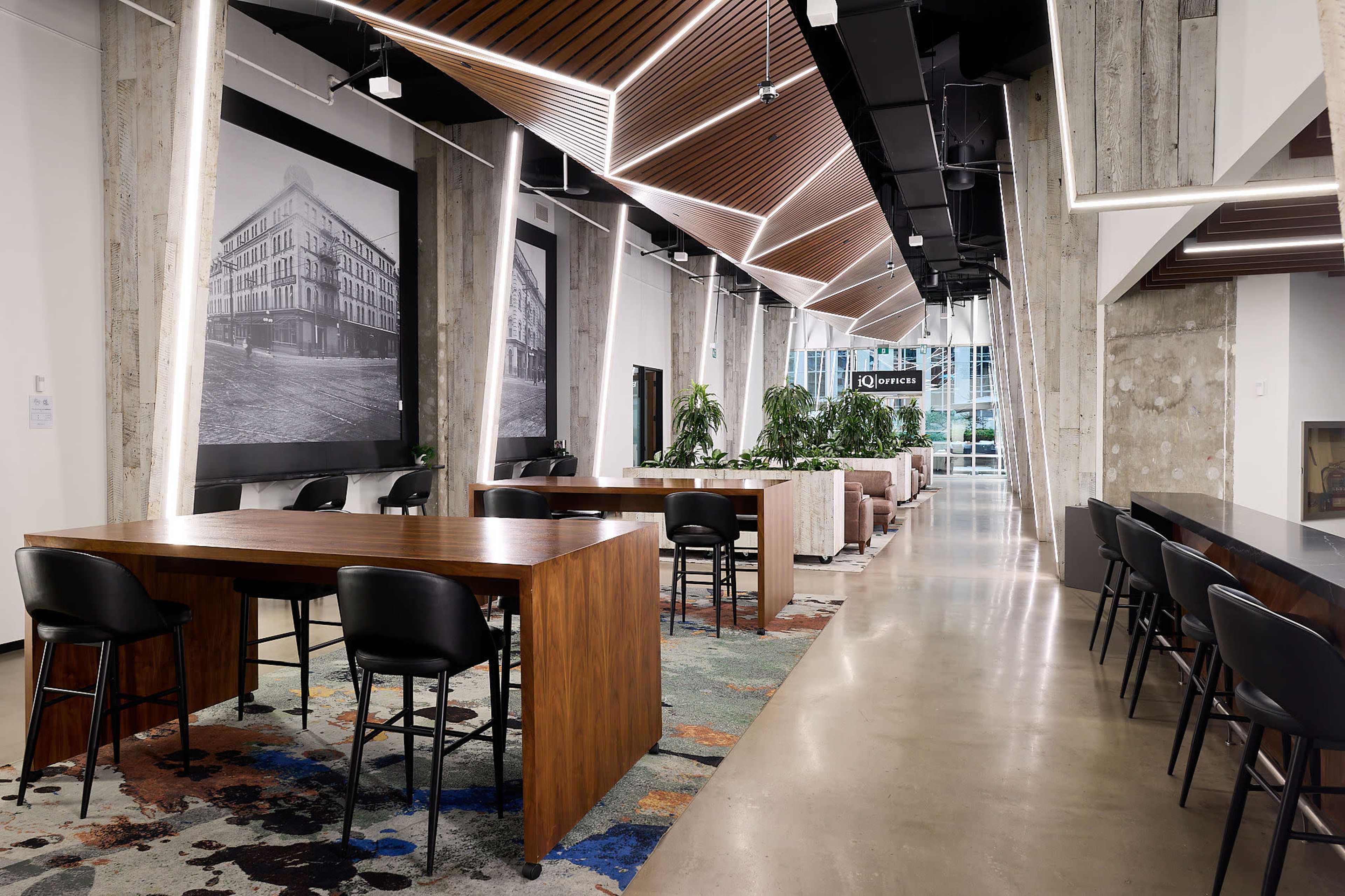 The image shows a modern, open workspace featuring wooden tables, black chairs, and large photos on the walls, with a polished concrete floor and greenery throughout.