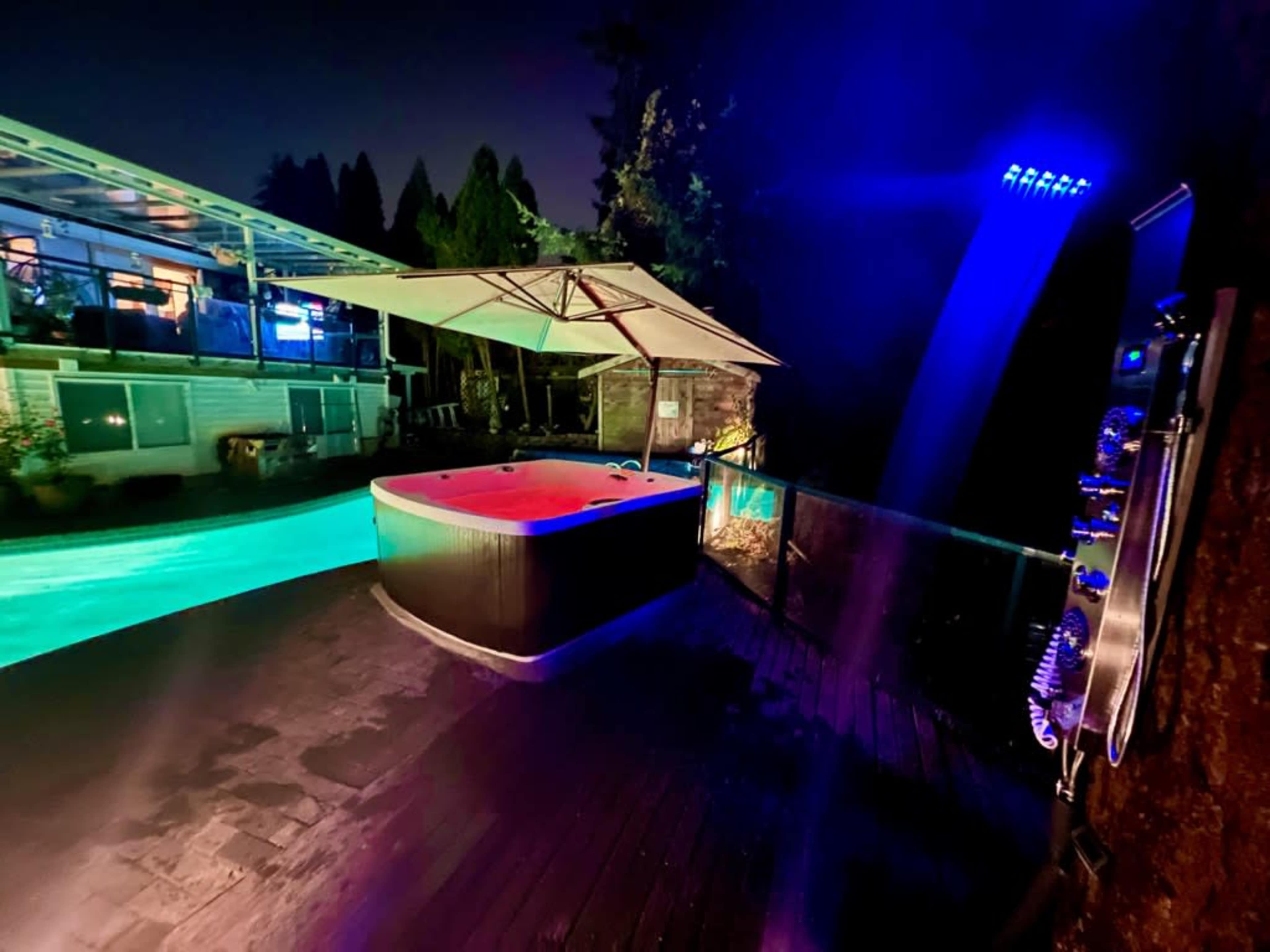 A hot tub with colorful lighting on a wooden deck next to a swimming pool, surrounded by trees at night.