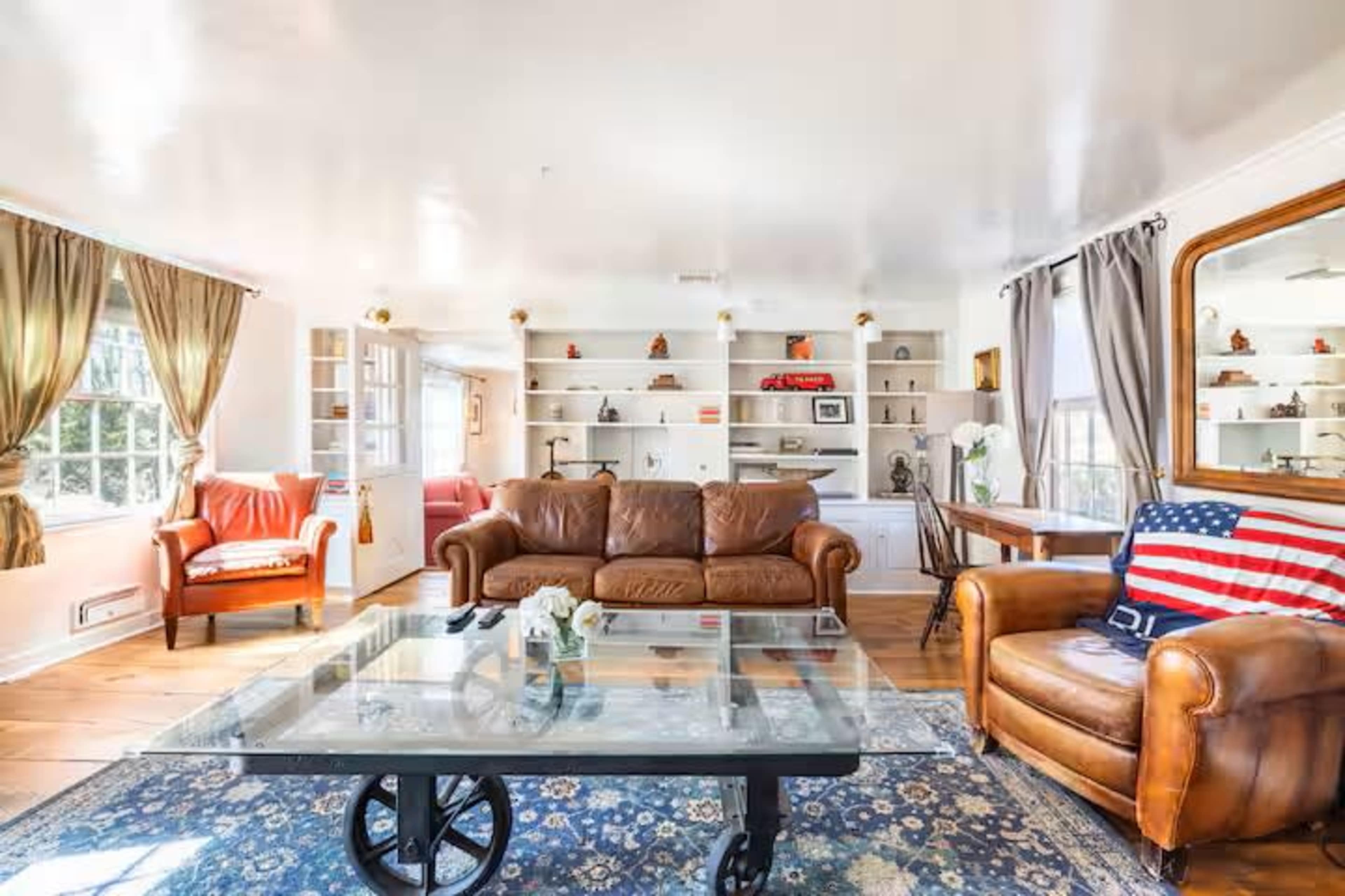 The image depicts a bright living room featuring a brown leather couch, an American flag throw pillow, and shelves displaying various decorative items.