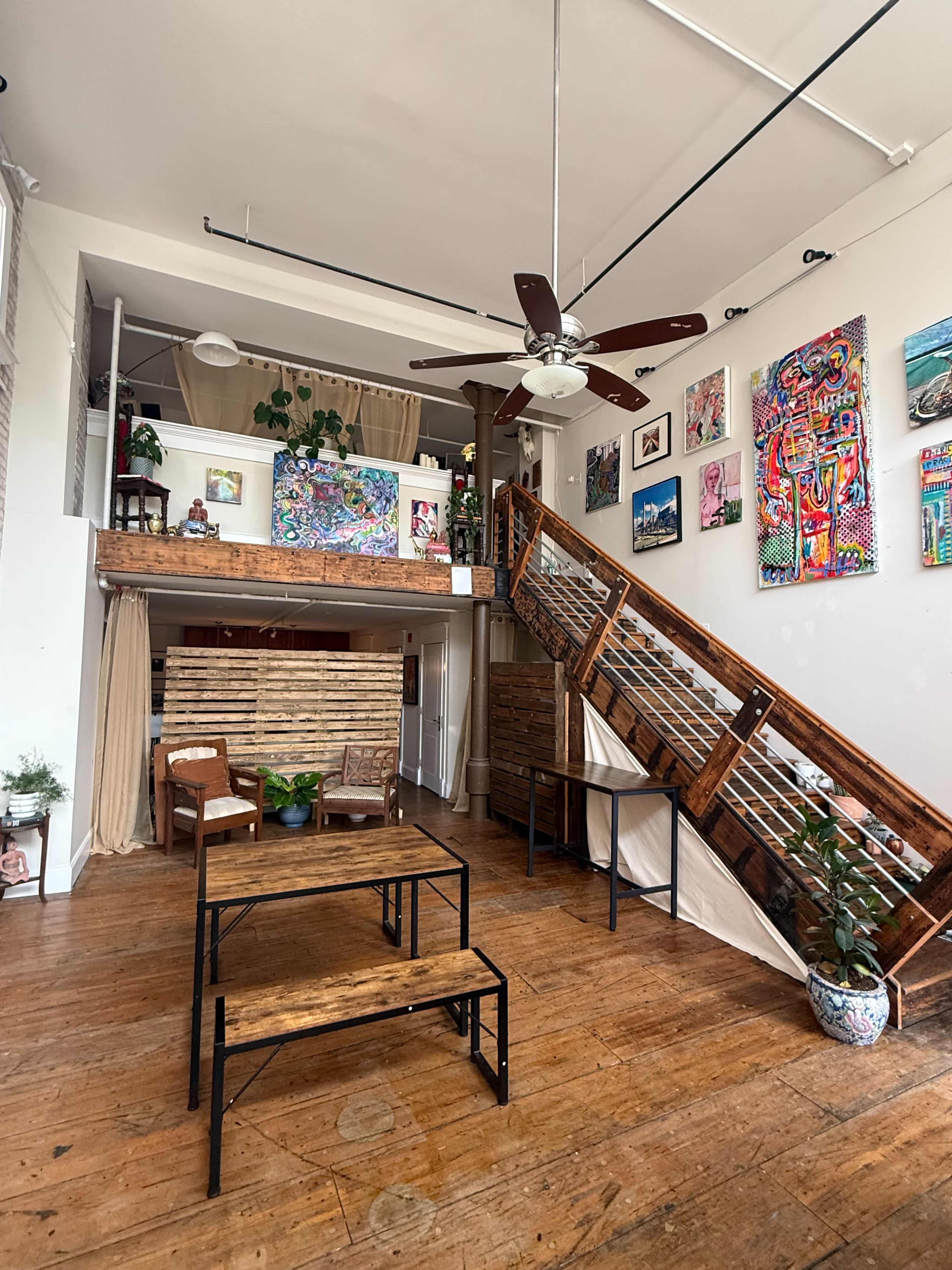 The image shows a cozy interior space featuring a wooden staircase, a small seating area with chairs, and vibrant artwork displayed on the walls.