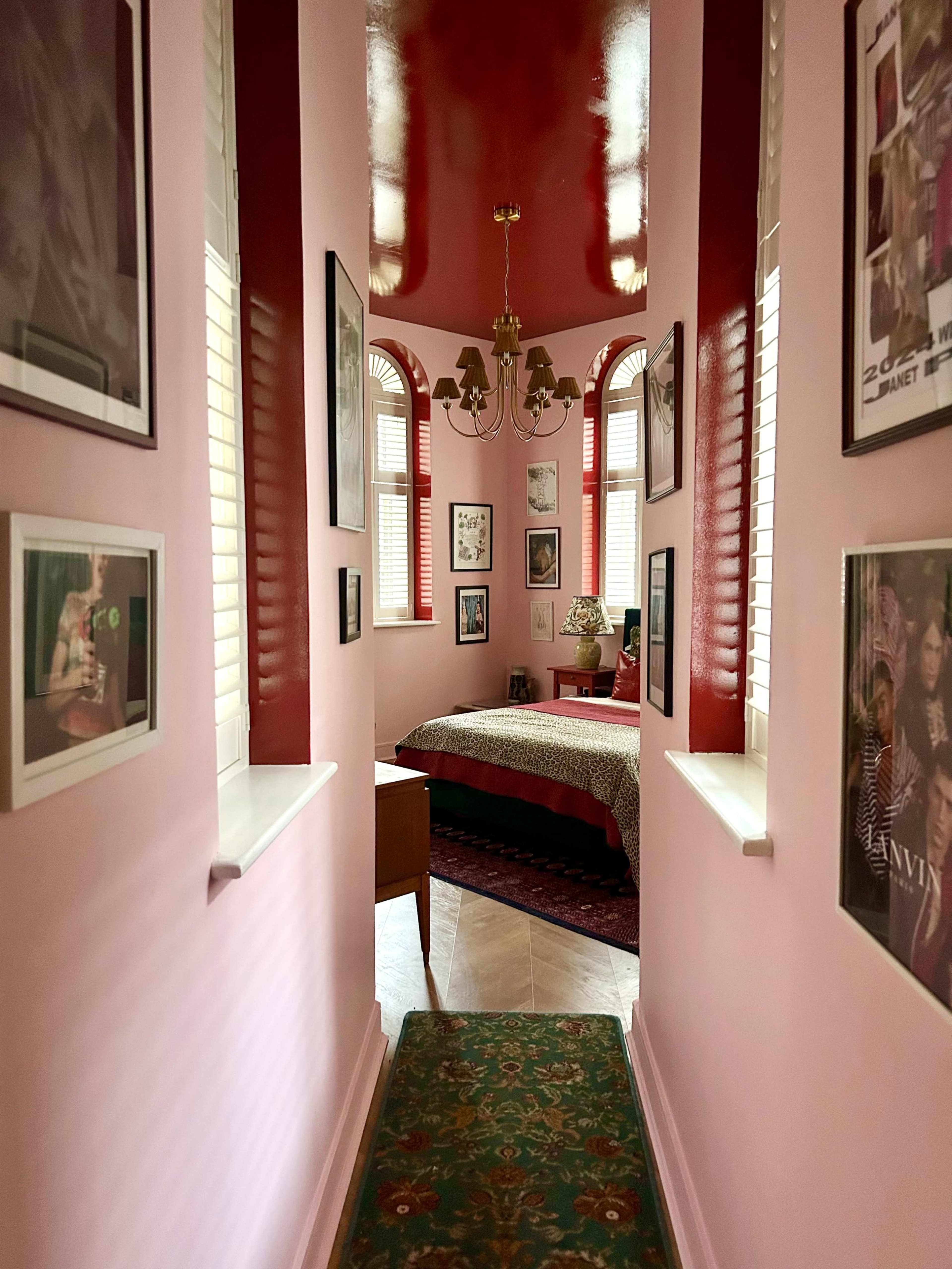 The image shows a narrow hallway lined with framed photographs leading to a bedroom with a patterned bedspread and decorative items.