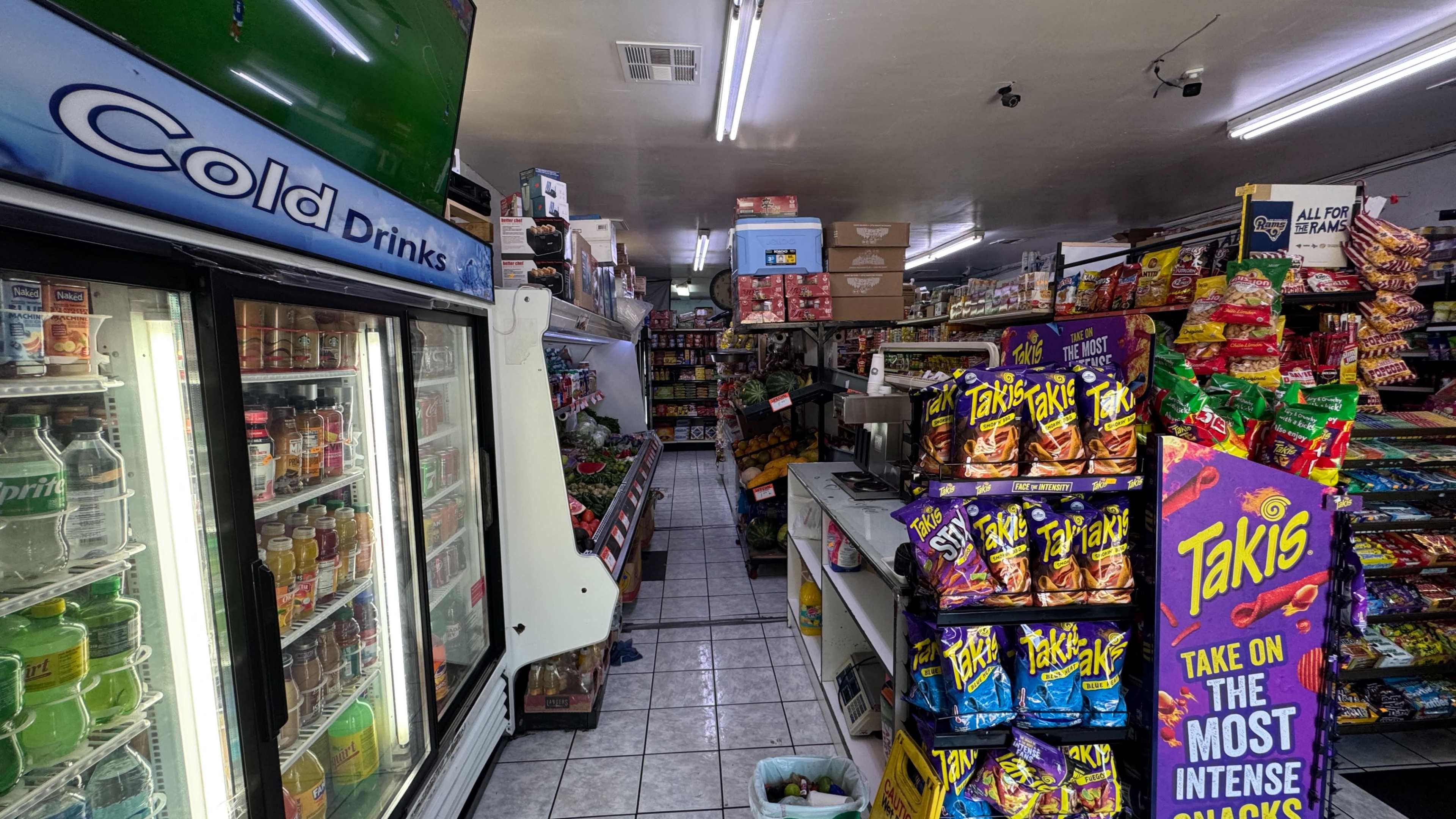 The image shows the interior of a small convenience store with aisles stocked with snacks, beverages, and various groceries.