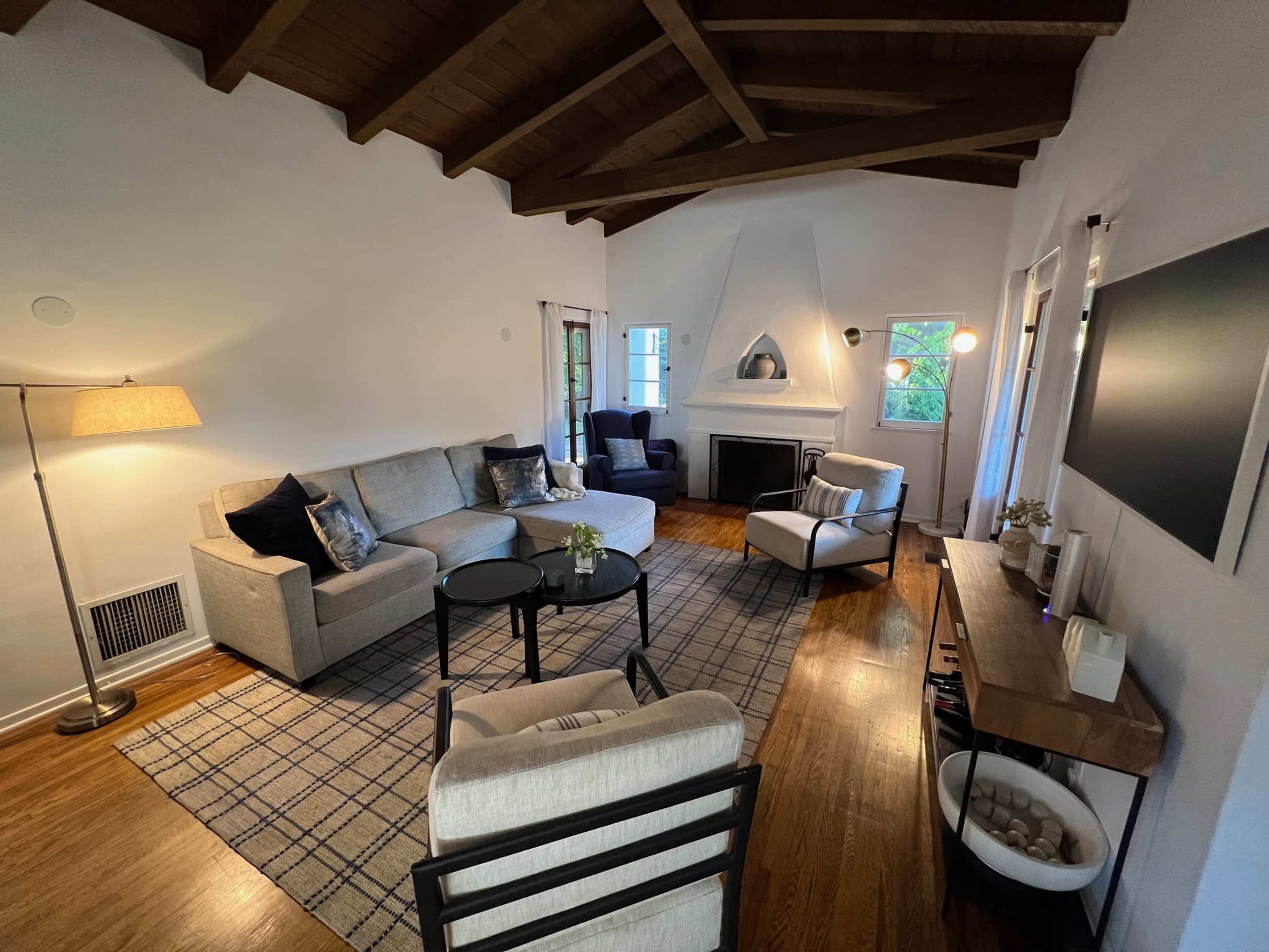 The image shows a living room with a sofa, armchairs, a coffee table, and a fireplace, all arranged under a ceiling with exposed wooden beams.