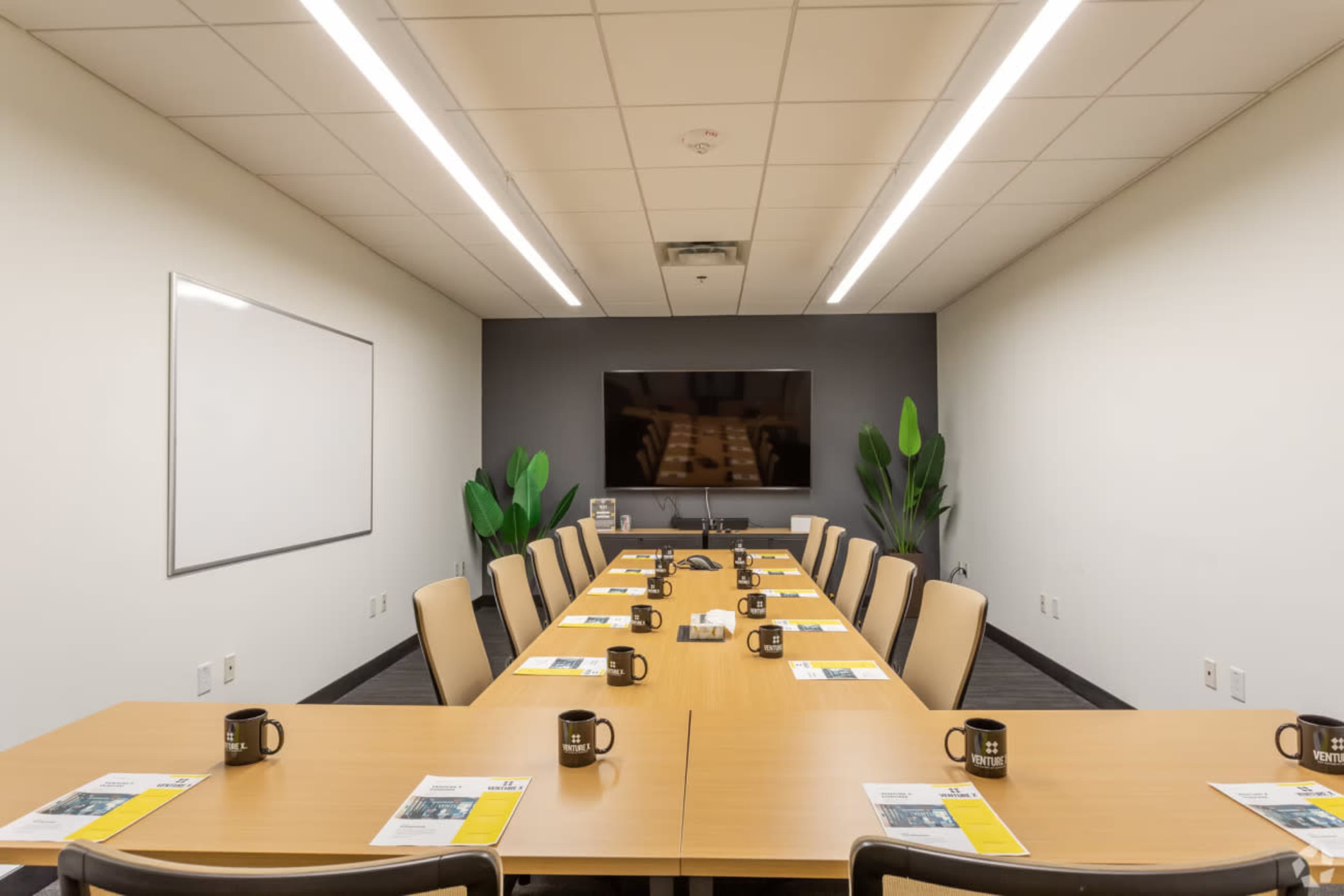 A conference room table is set up for a meeting, with place settings, mugs, and a large screen at one end.