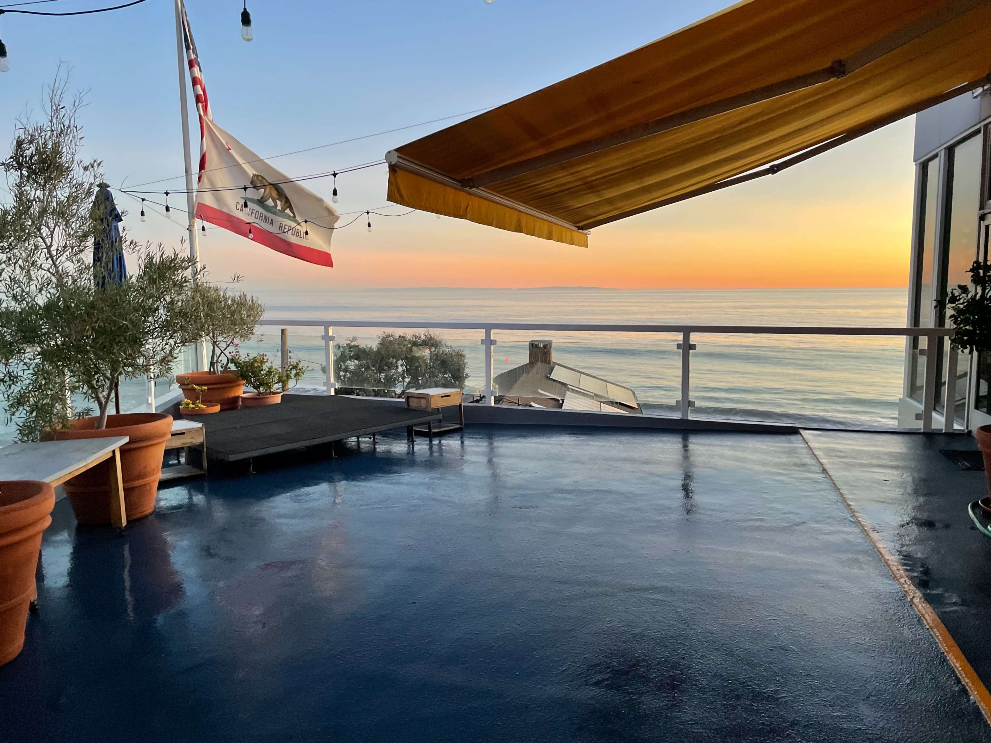 A patio with potted plants and a California flag overlooks a calm ocean at sunset.