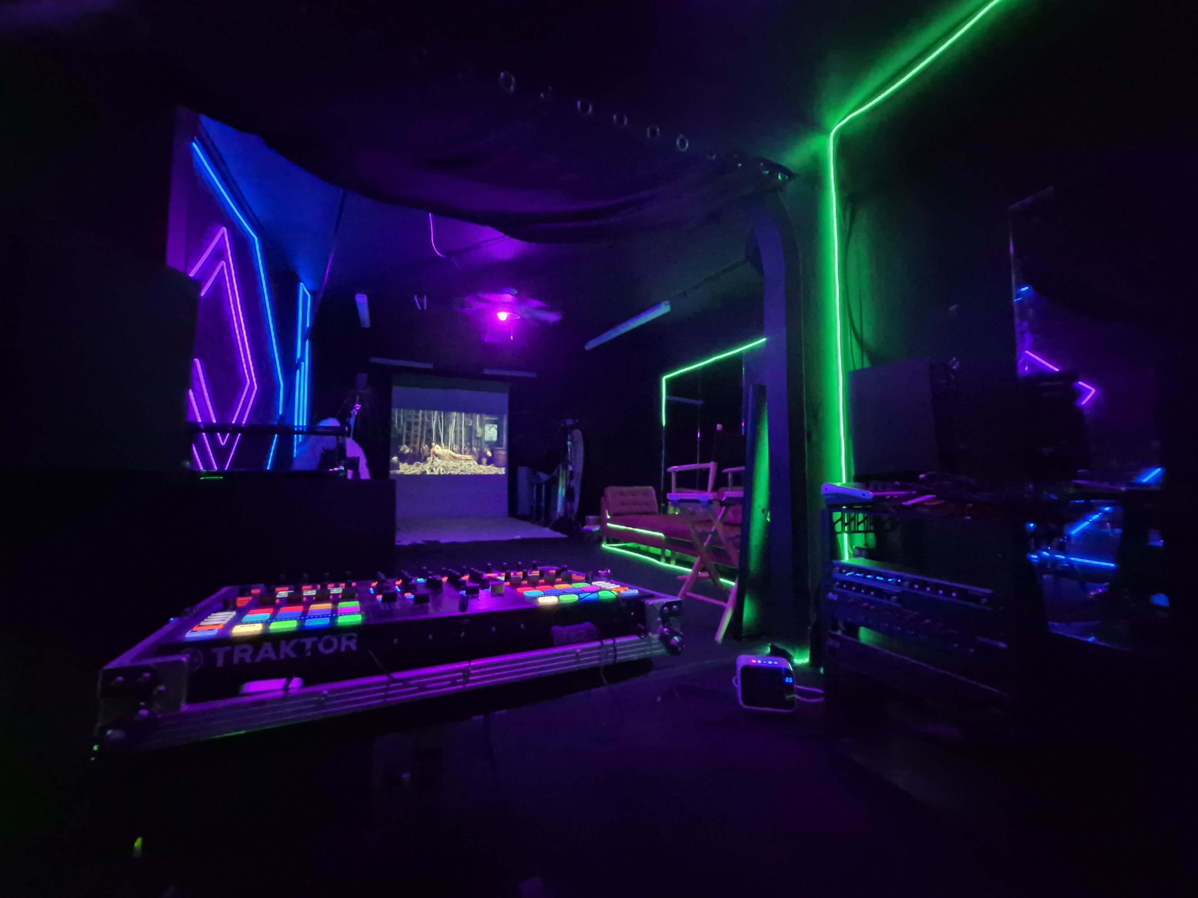 A dimly lit room features colorful neon lights and a projection screen showcasing a nature scene, with a DJ controller prominently displayed in the foreground.