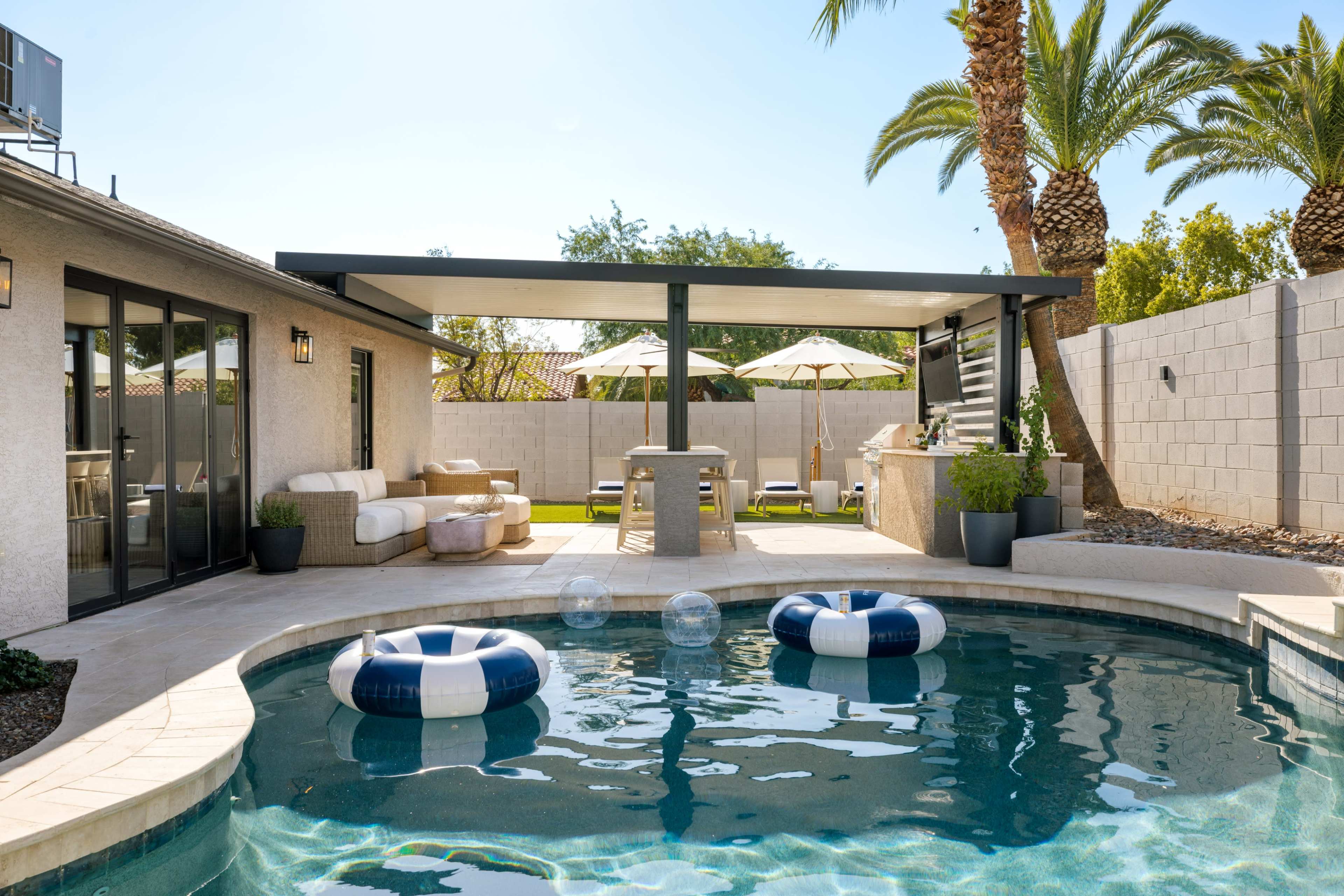 The image shows a modern pool area featuring a swimming pool with two inflatable floats, surrounded by palm trees and a covered patio with seating and umbrellas.