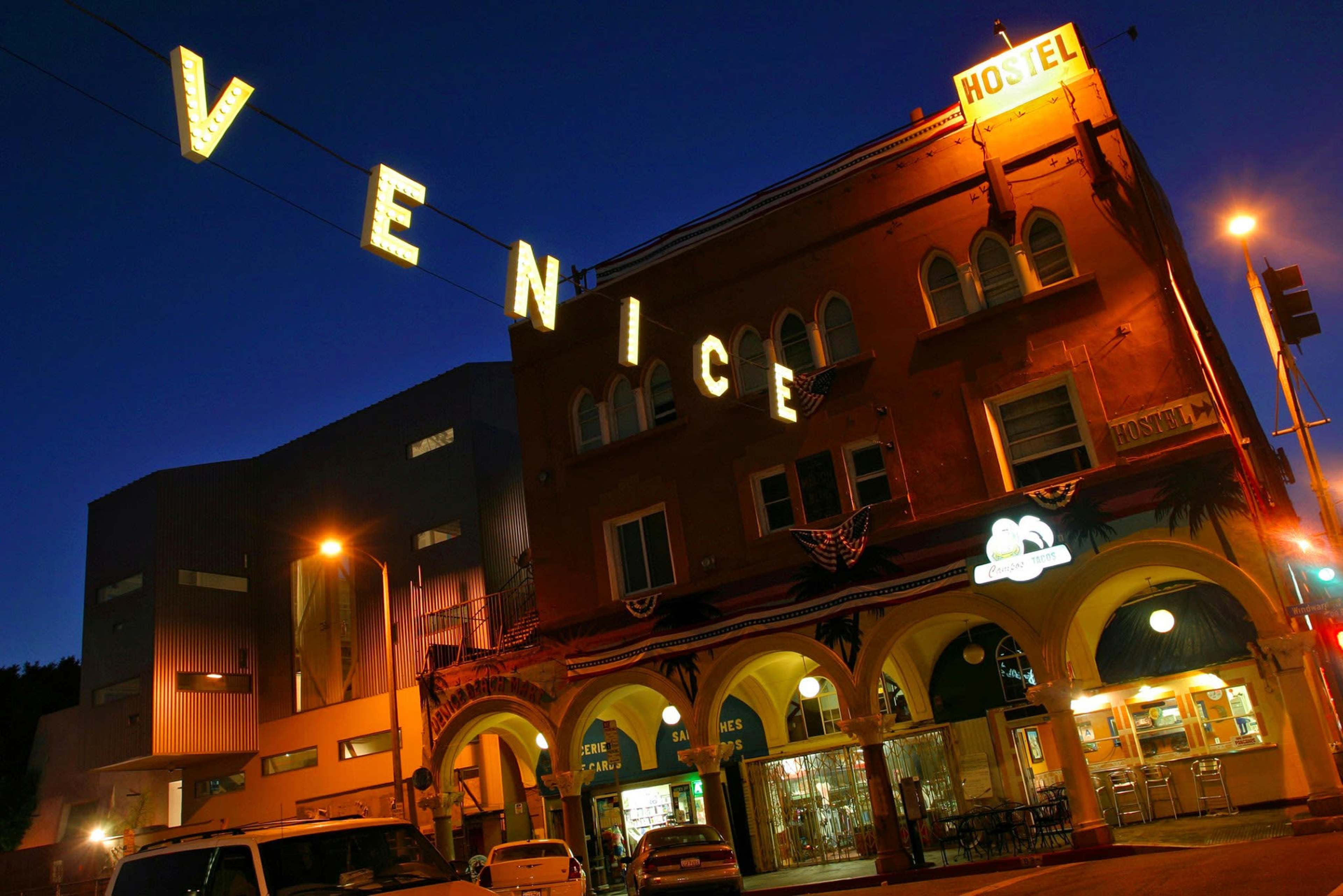 A brightly lit hostel with "VENICE" signage is seen at night, located in a building featuring arched entrances and decorative elements.