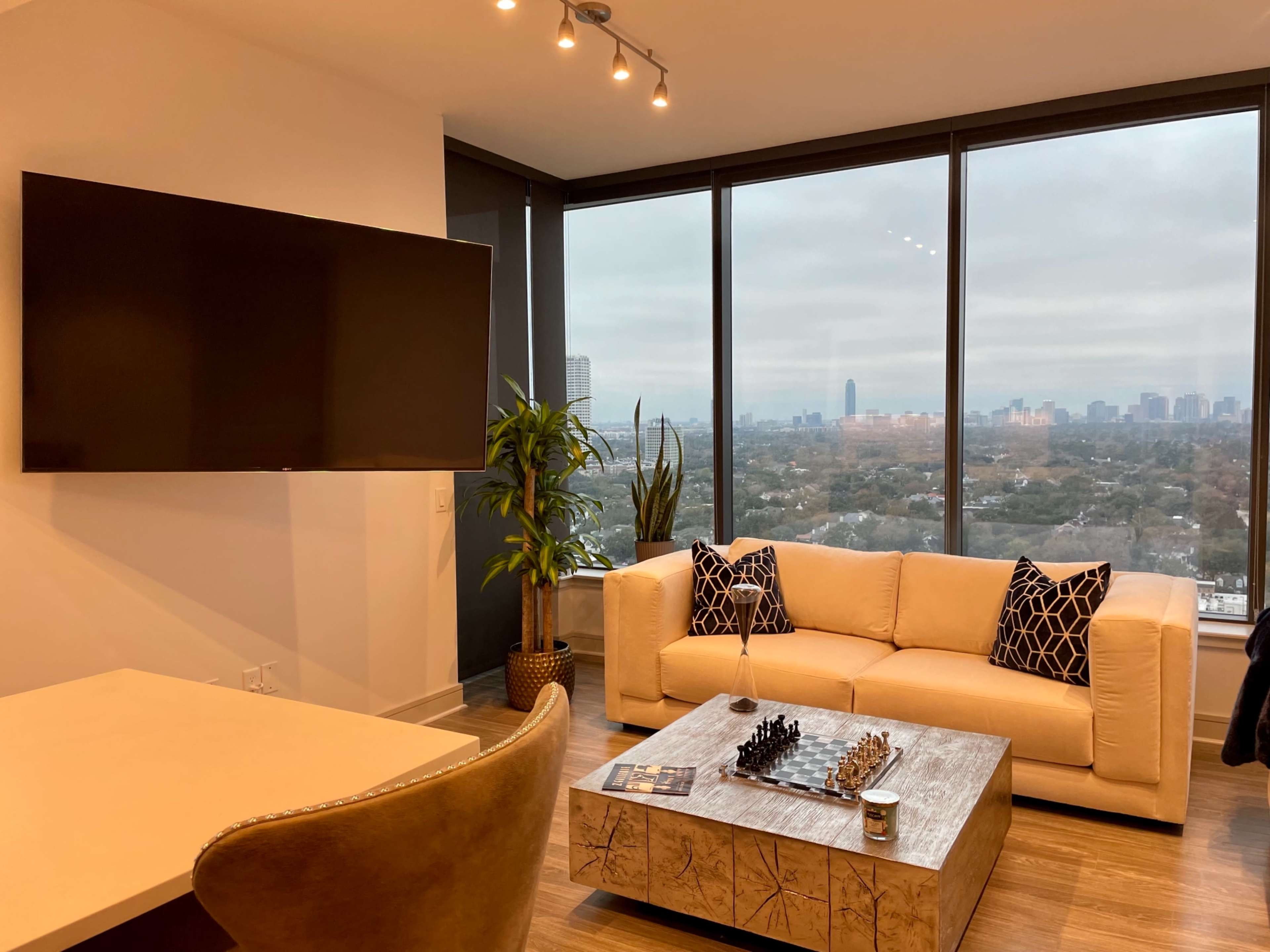 A modern living room features a beige sofa with patterned cushions, a coffee table with a chess set, and large windows offering a city view.