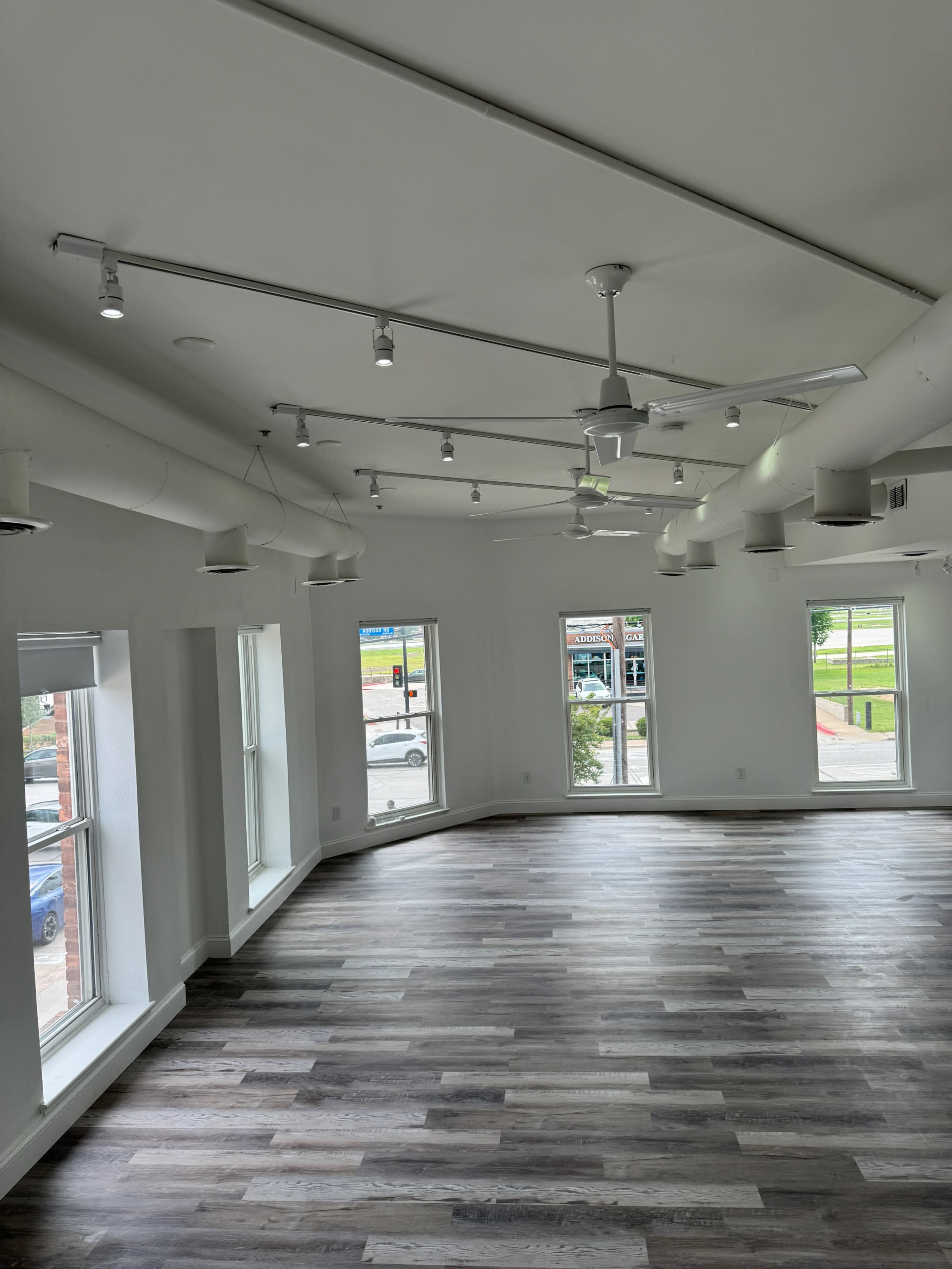 The image shows a bright, empty room with large windows, a wooden floor, and multiple ceiling fans and light fixtures.