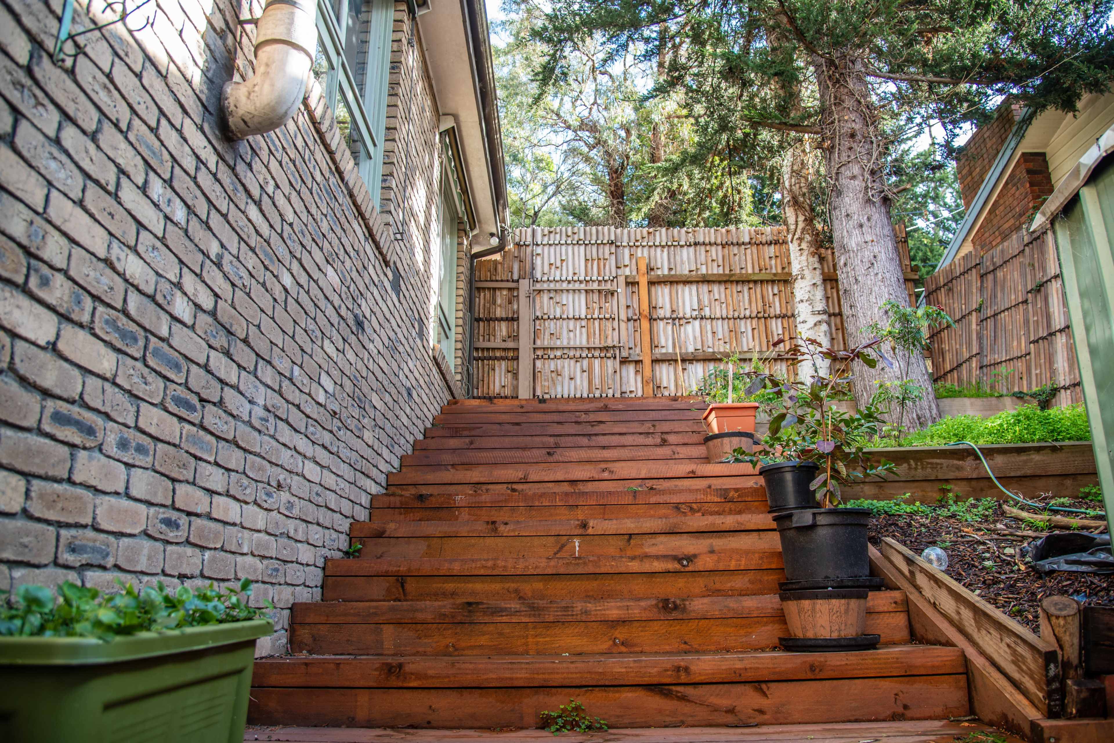 The image shows a wooden staircase leading up to a fenced area surrounded by greenery and a brick wall.