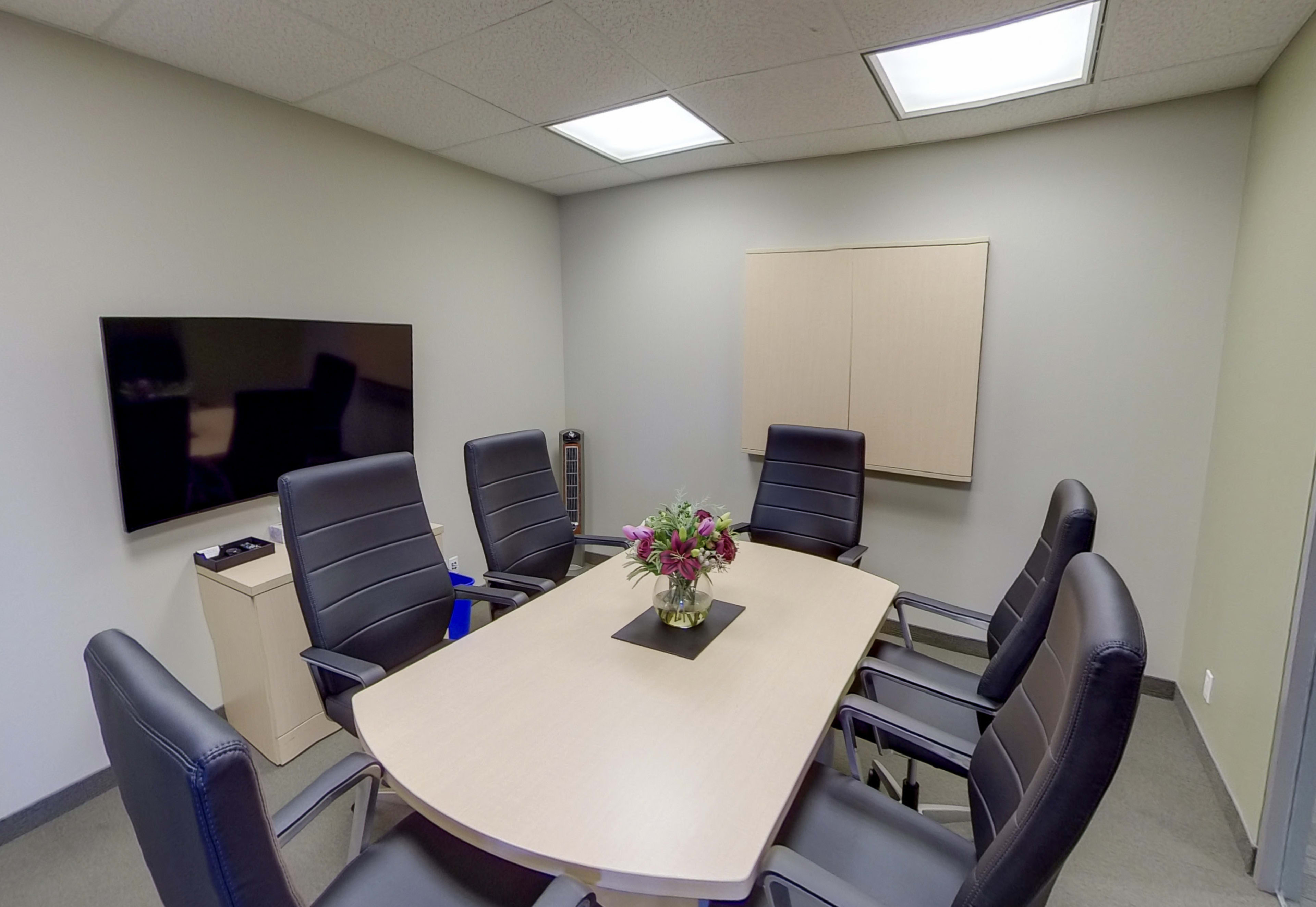 A conference room is set up with a large table surrounded by six black chairs, a wall-mounted screen, and a flower arrangement in the center.