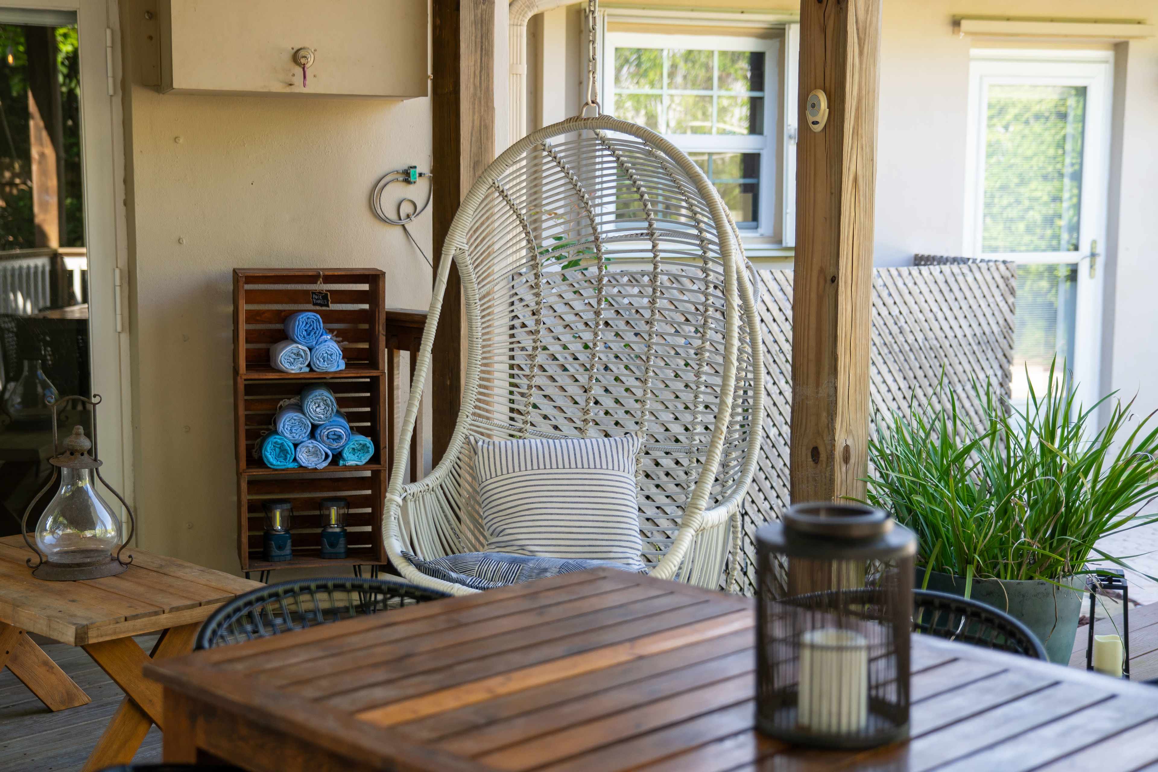 A hanging chair is positioned on a patio next to a wooden table, with neatly stacked towels and plants in the background.