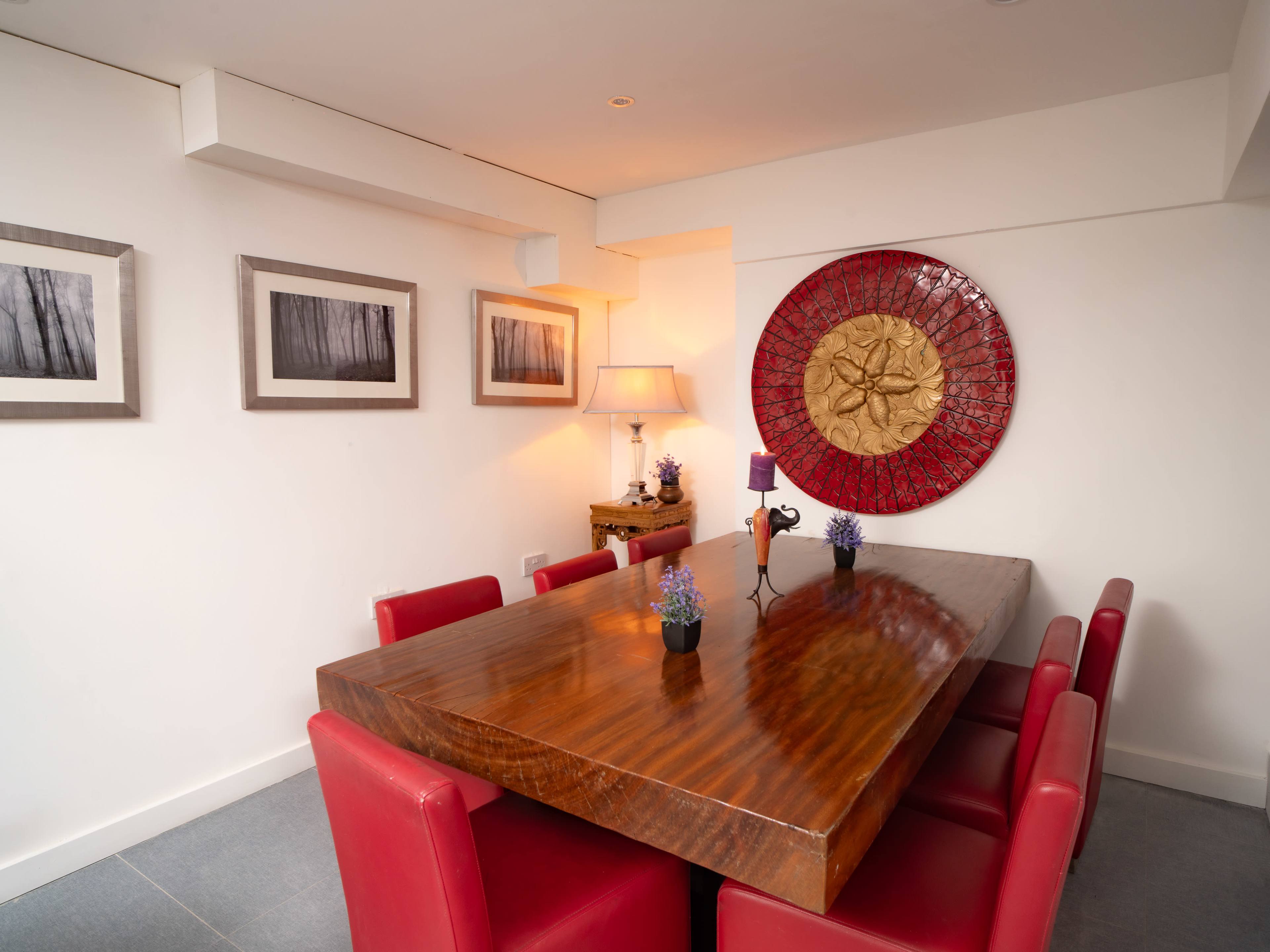A large wooden dining table surrounded by red chairs is set in a minimalist room with framed photographs on the walls and a decorative wall piece.