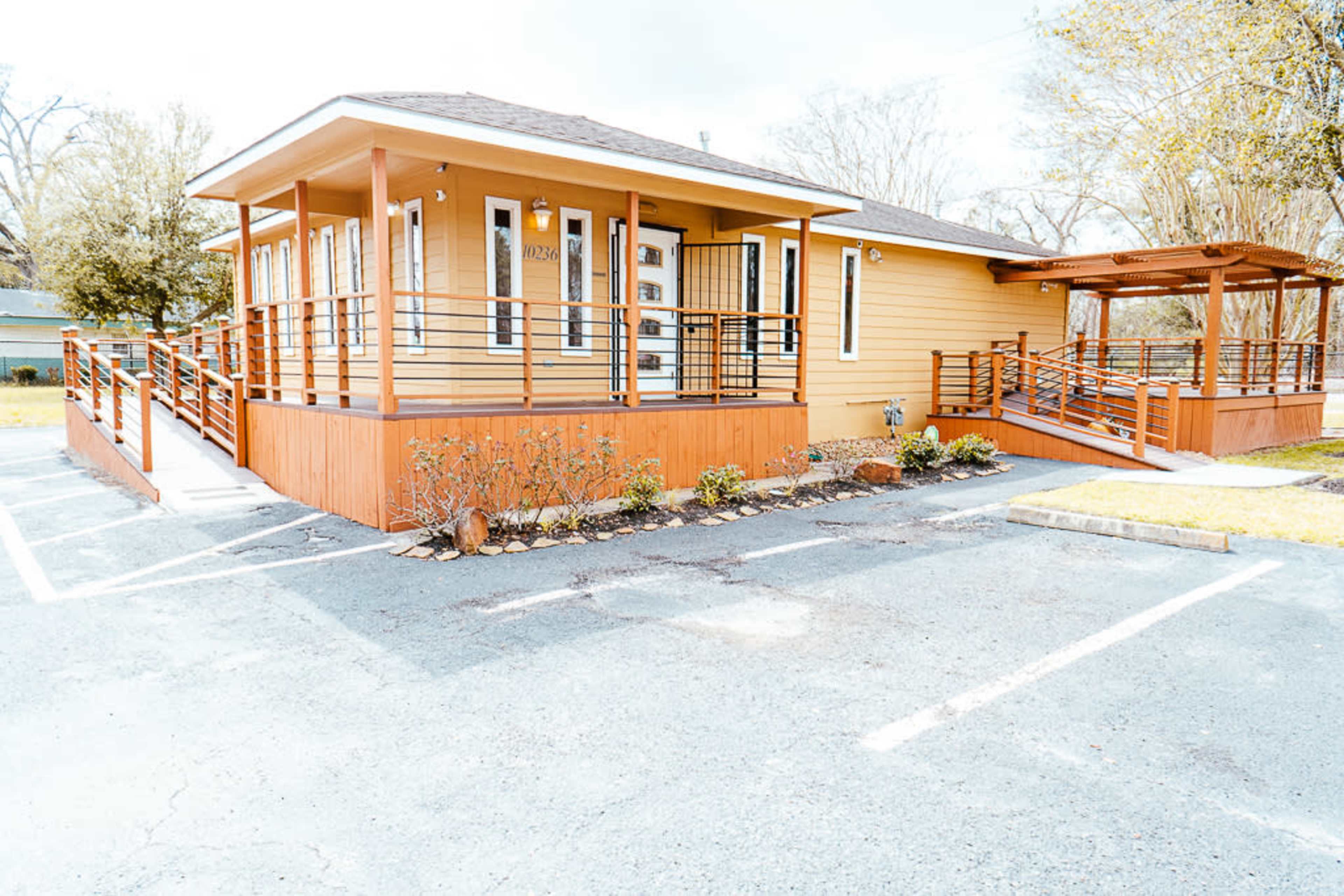 A yellow wooden building with a ramp and porch in front, surrounded by landscaped greenery and a parking area.