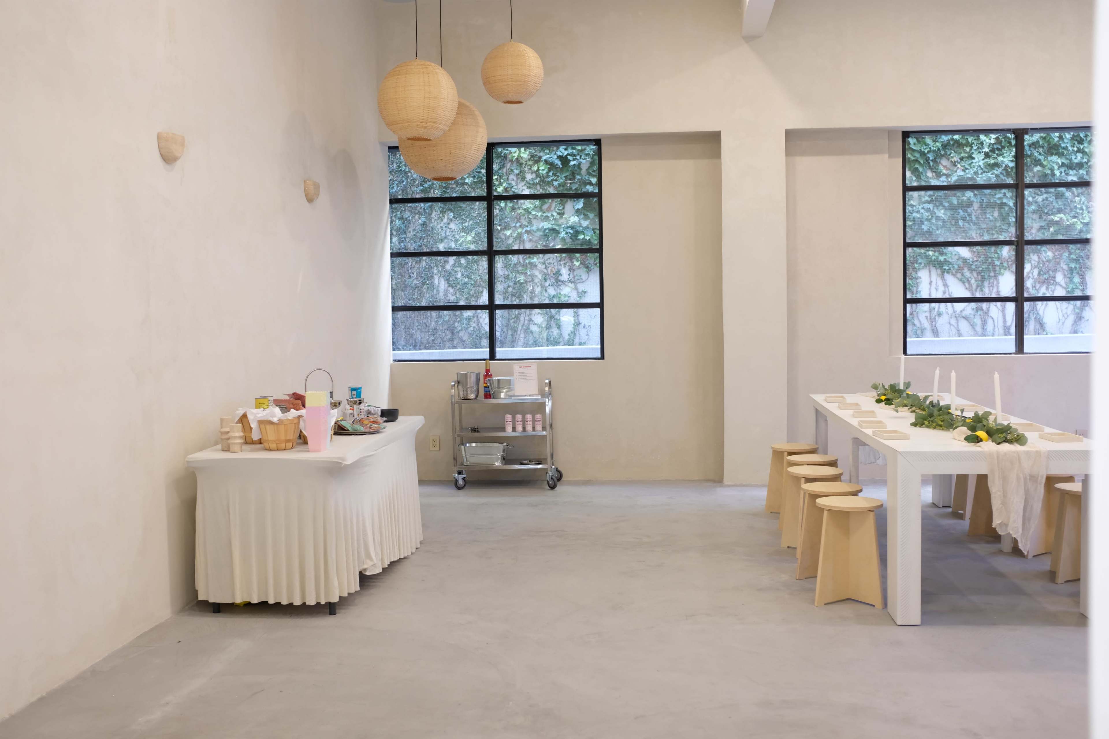 The image shows a minimalist interior space featuring a long dining table with stools, a serving cart, and decorative hanging lights.