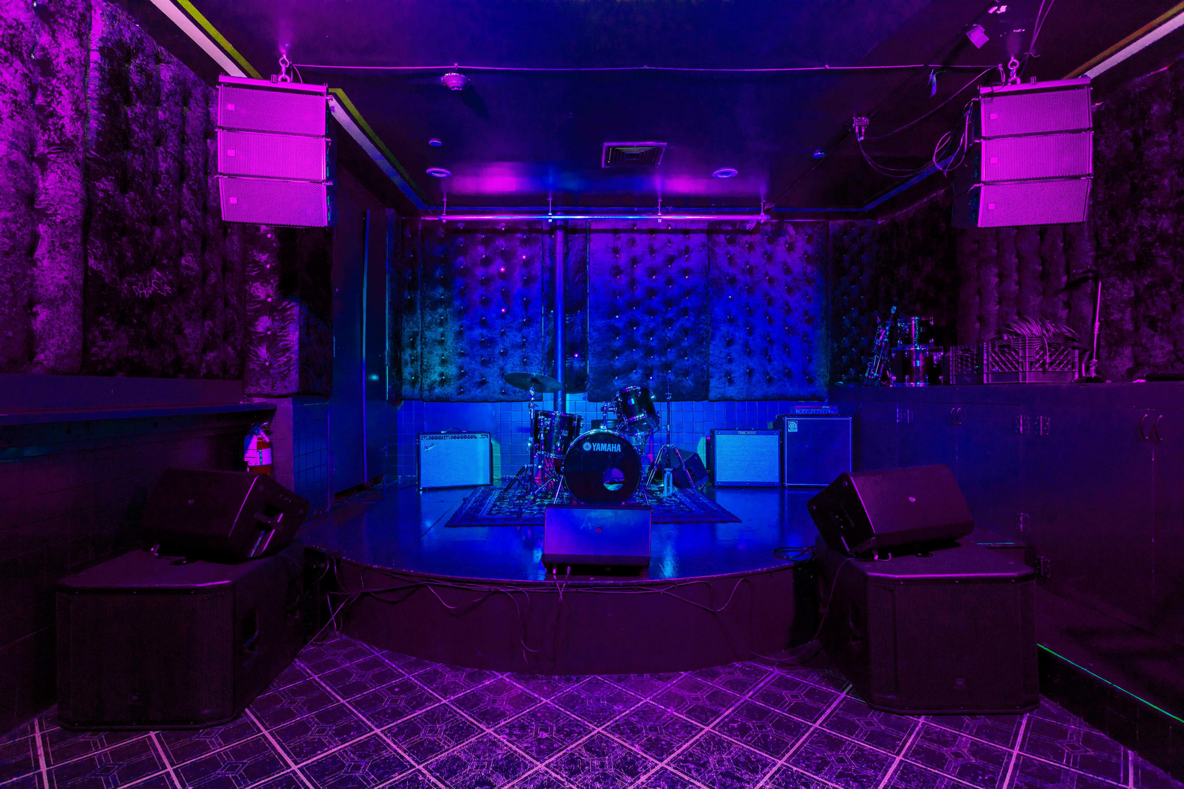 The image shows a dimly lit music stage with purple lighting, featuring a drum set and amplifiers on a raised platform.