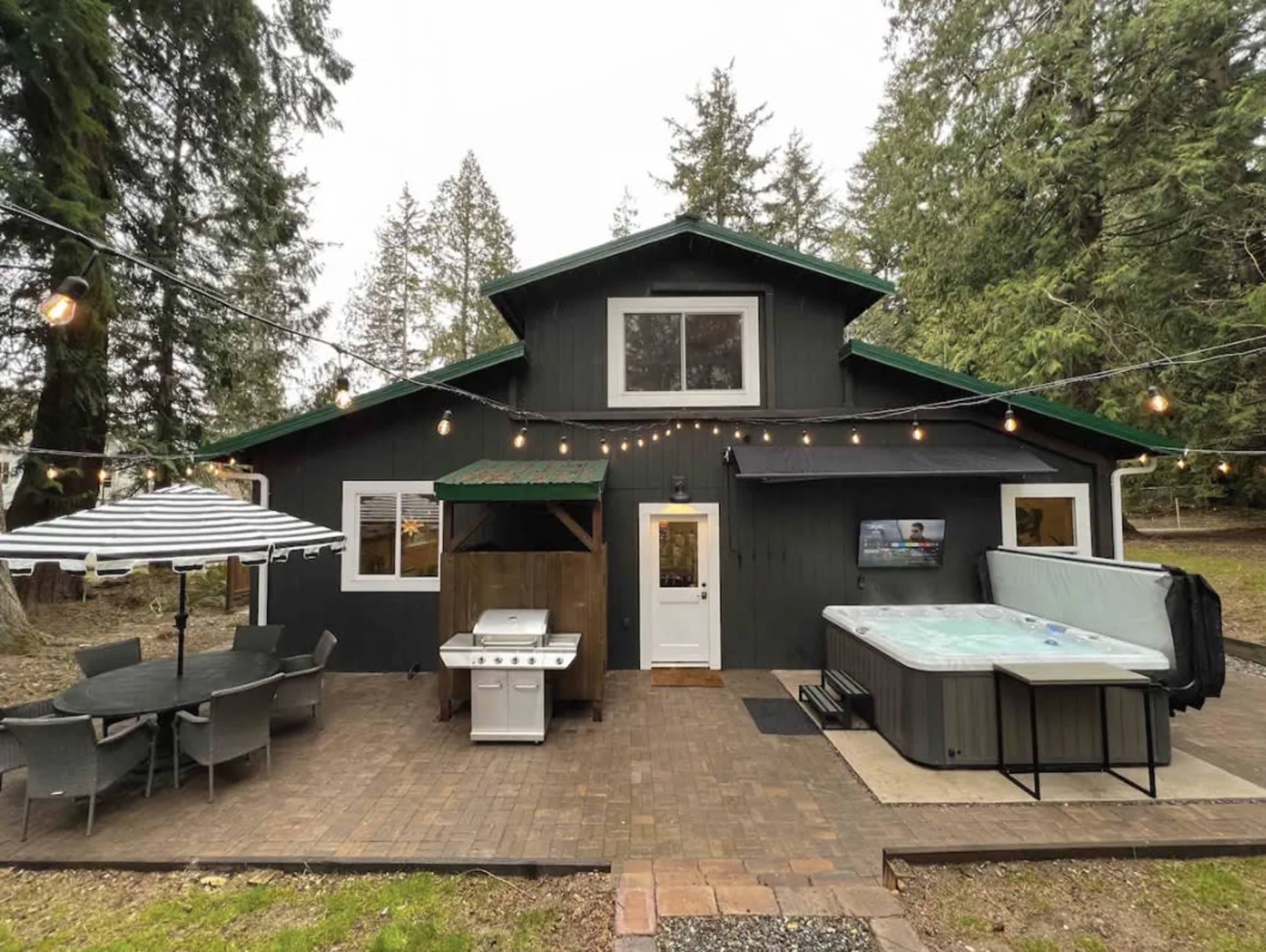 The image shows a cabin with a patio featuring a dining set, a grill, a hot tub, and outdoor string lights surrounded by trees.