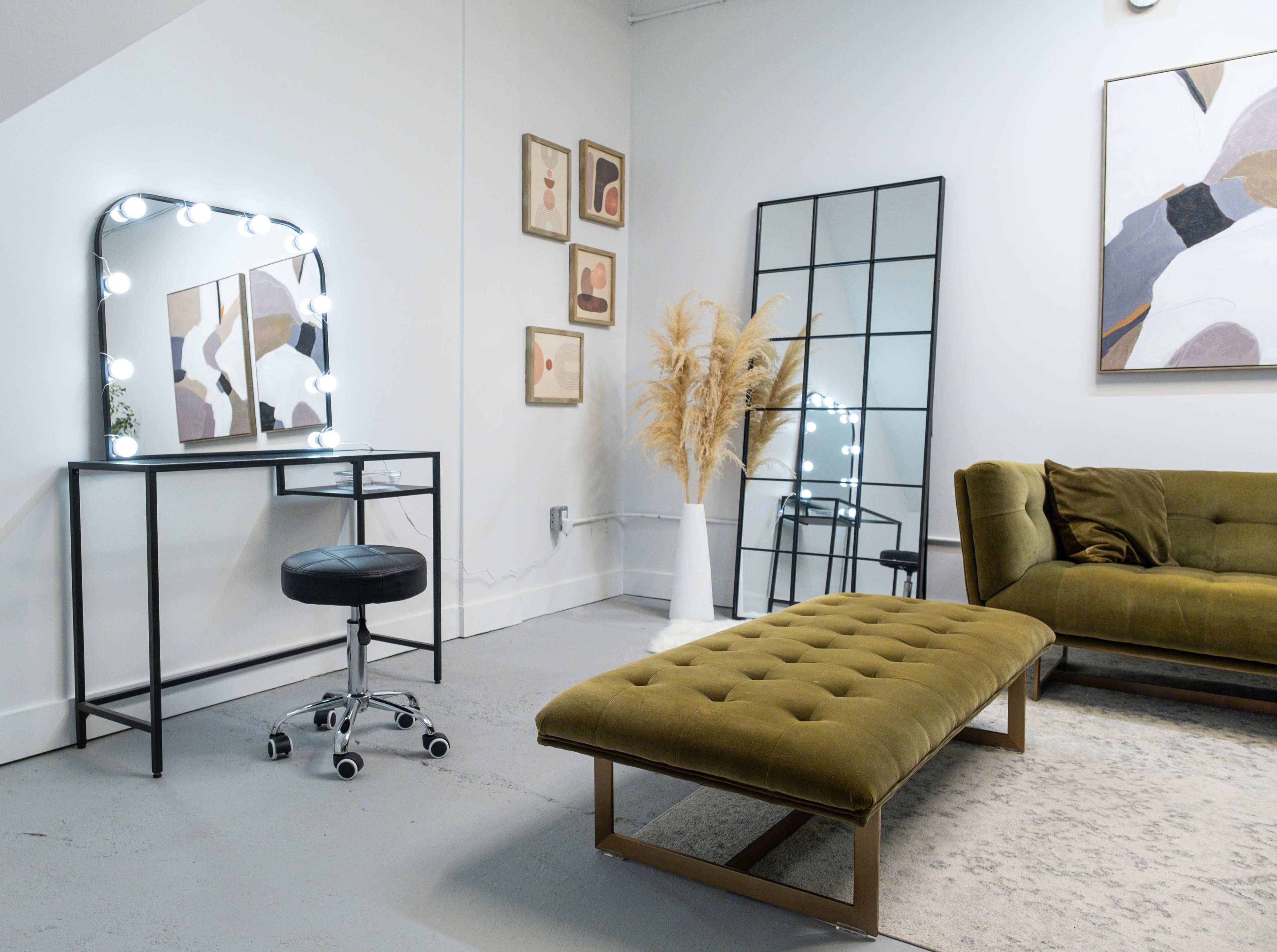 A modern interior scene features a green velvet sofa, a tufted bench, a mirrored vanity with lights, and decorative artwork on the walls.