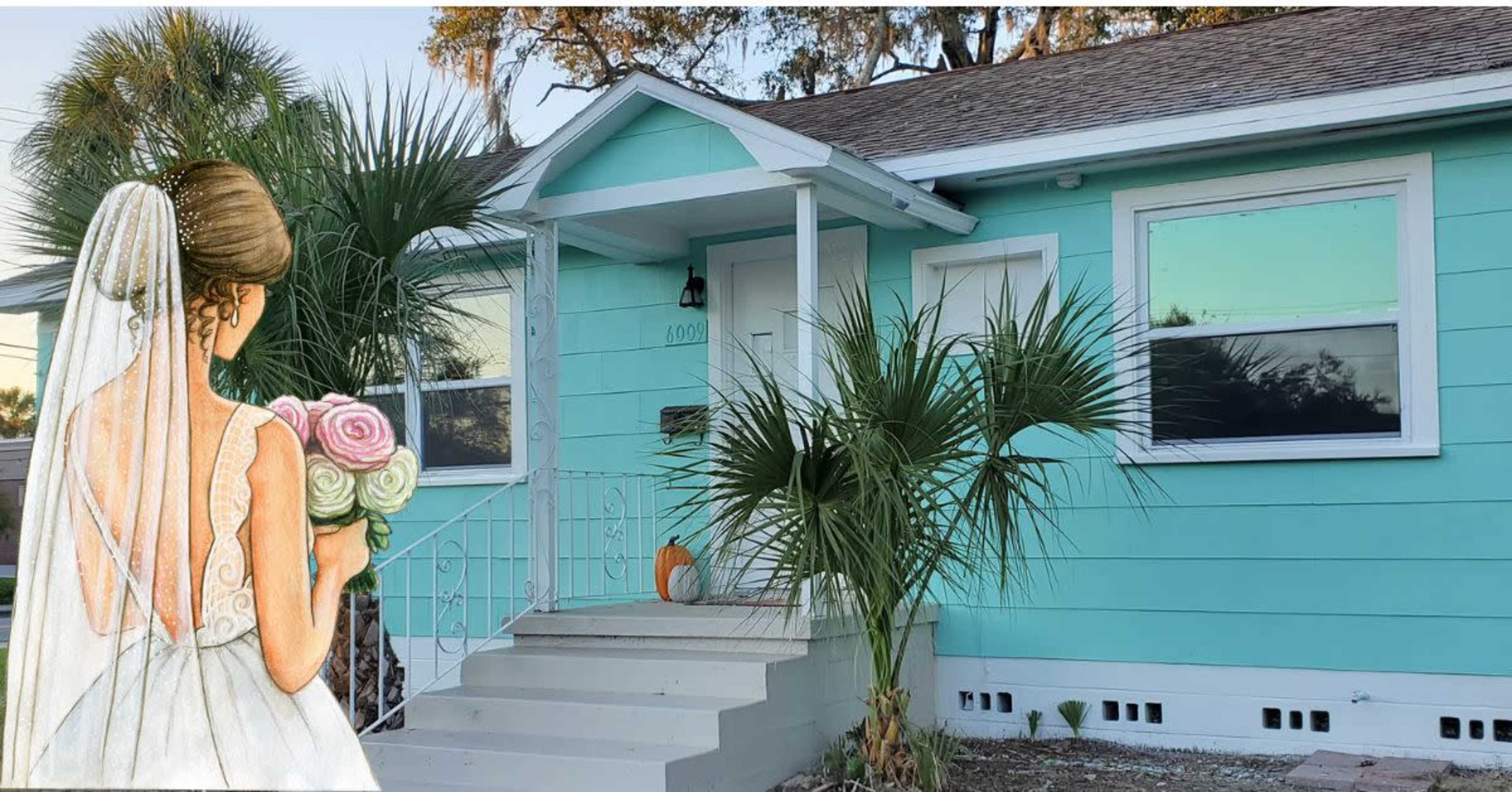 A bride holding a bouquet stands near the entrance of a turquoise house with palm trees in the front.