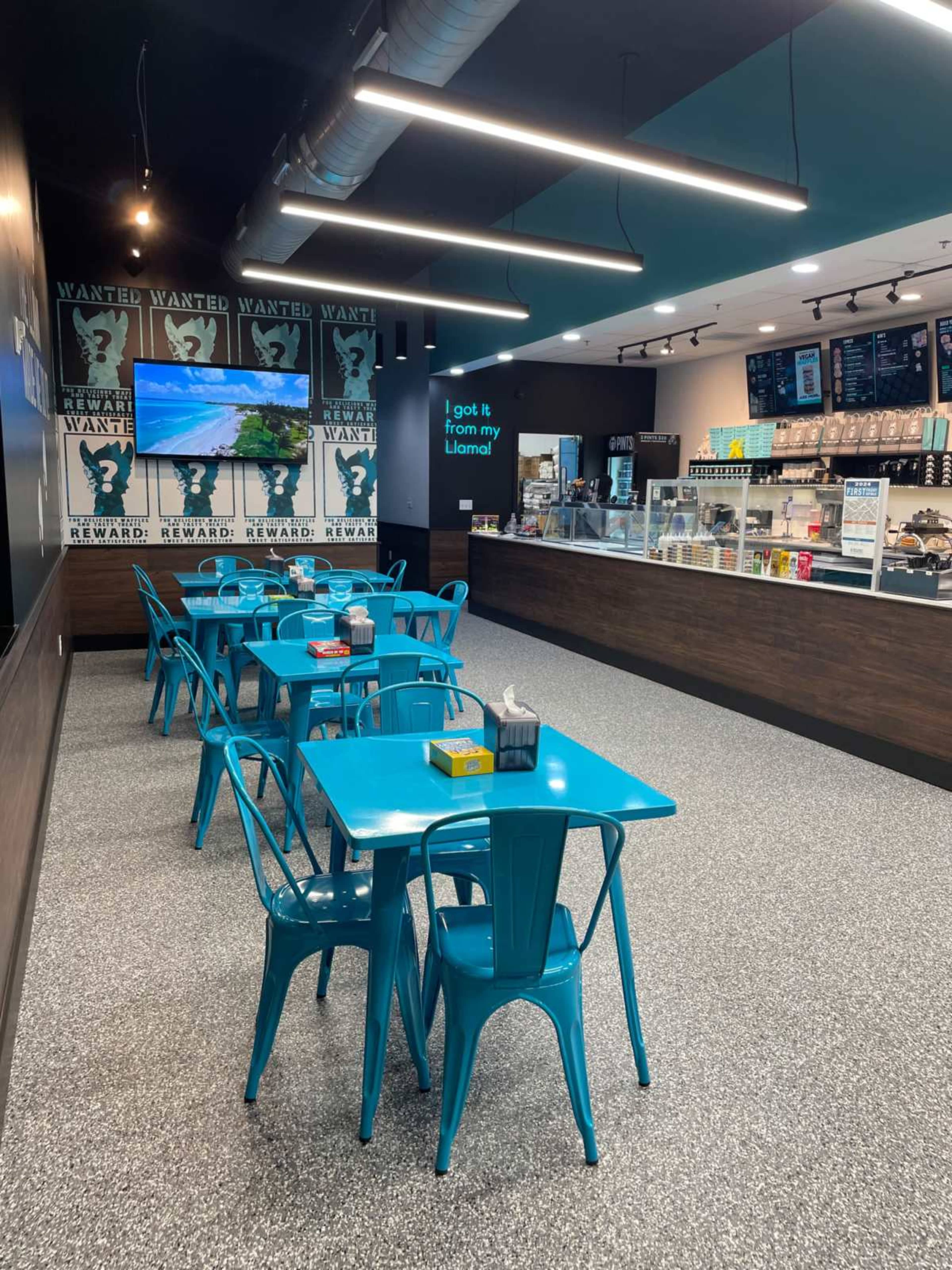 A brightly lit café features turquoise tables and chairs arranged on a speckled gray floor, with a serving counter and a wall decorated with wanted posters.