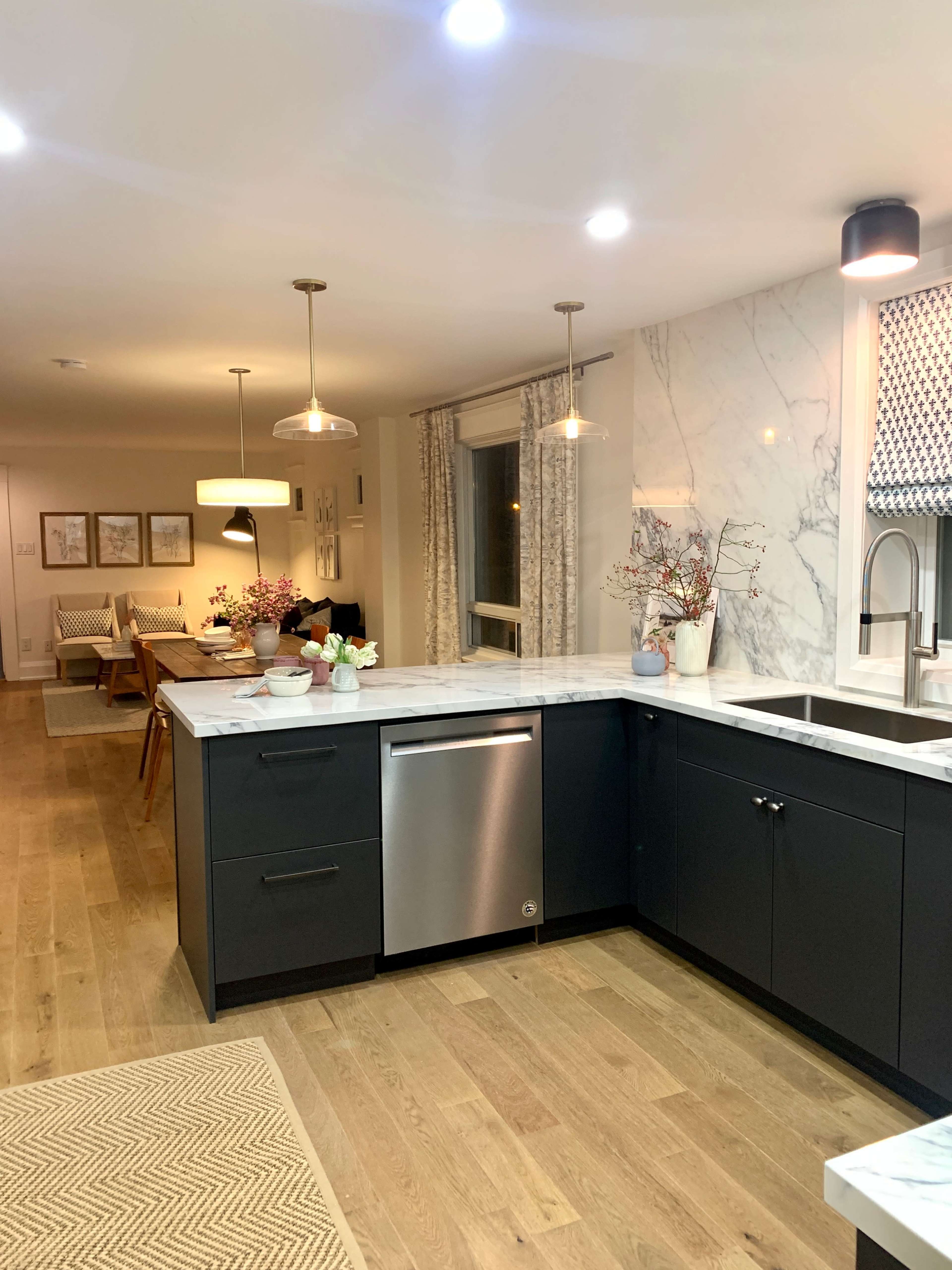 A modern kitchen features dark cabinetry, a stainless steel dishwasher, and a marble countertop, with a dining area visible in the background.