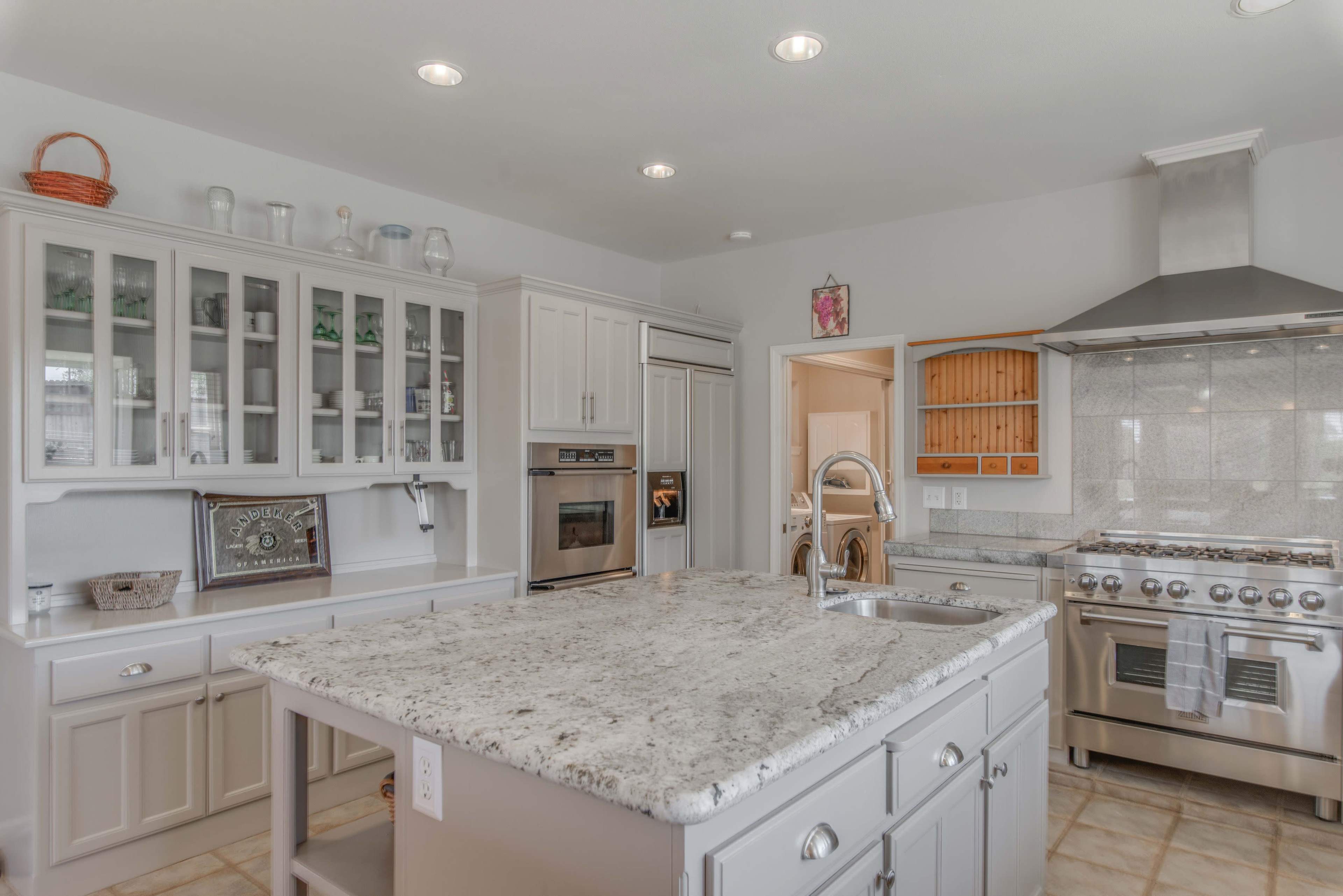 A modern kitchen features a large granite island, stainless steel appliances, and glass-front cabinets.
