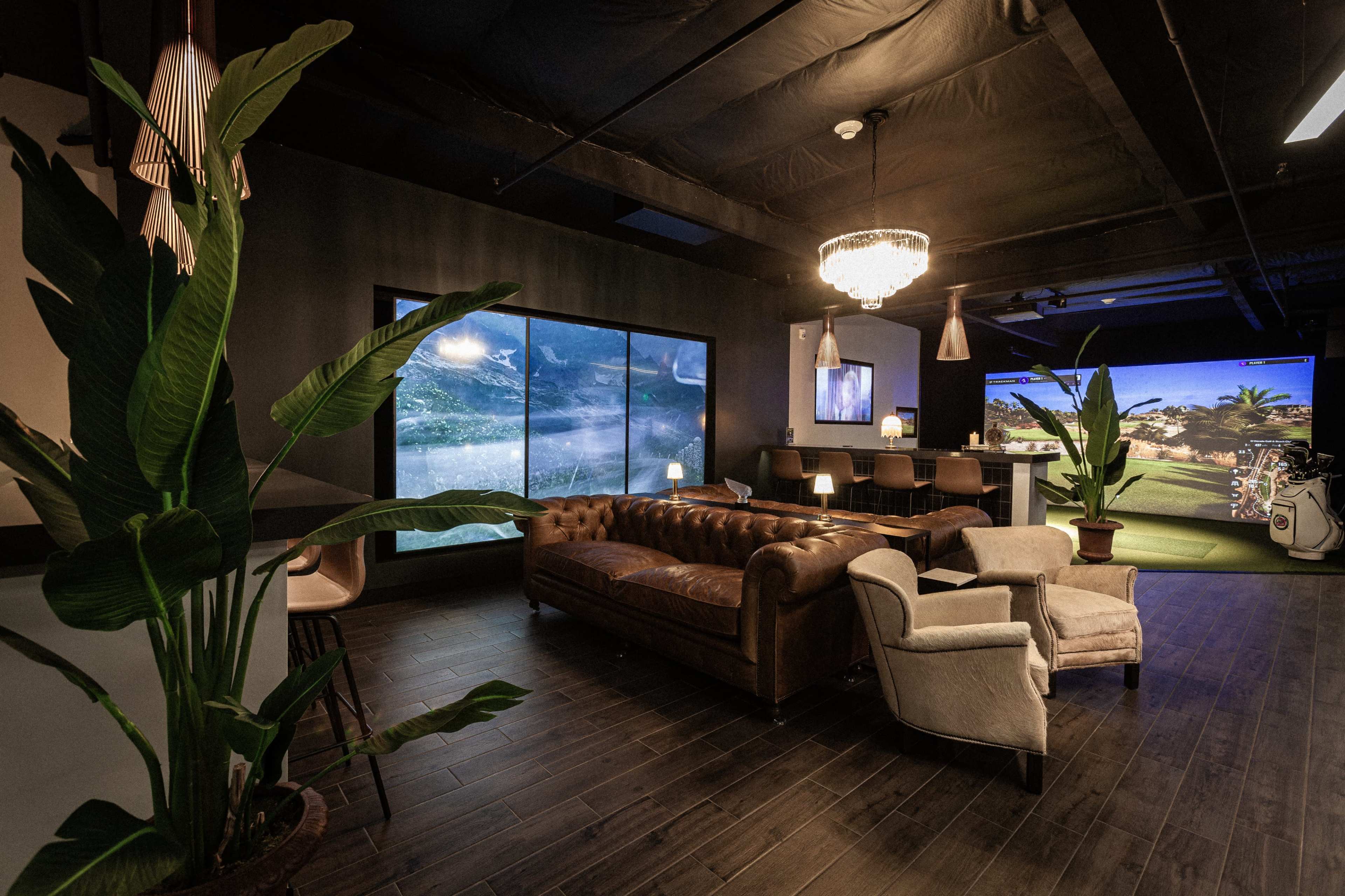 The image shows a modern indoor space featuring a leather sofa, armchairs, large windows, and golf simulation screens with tropical scenery.