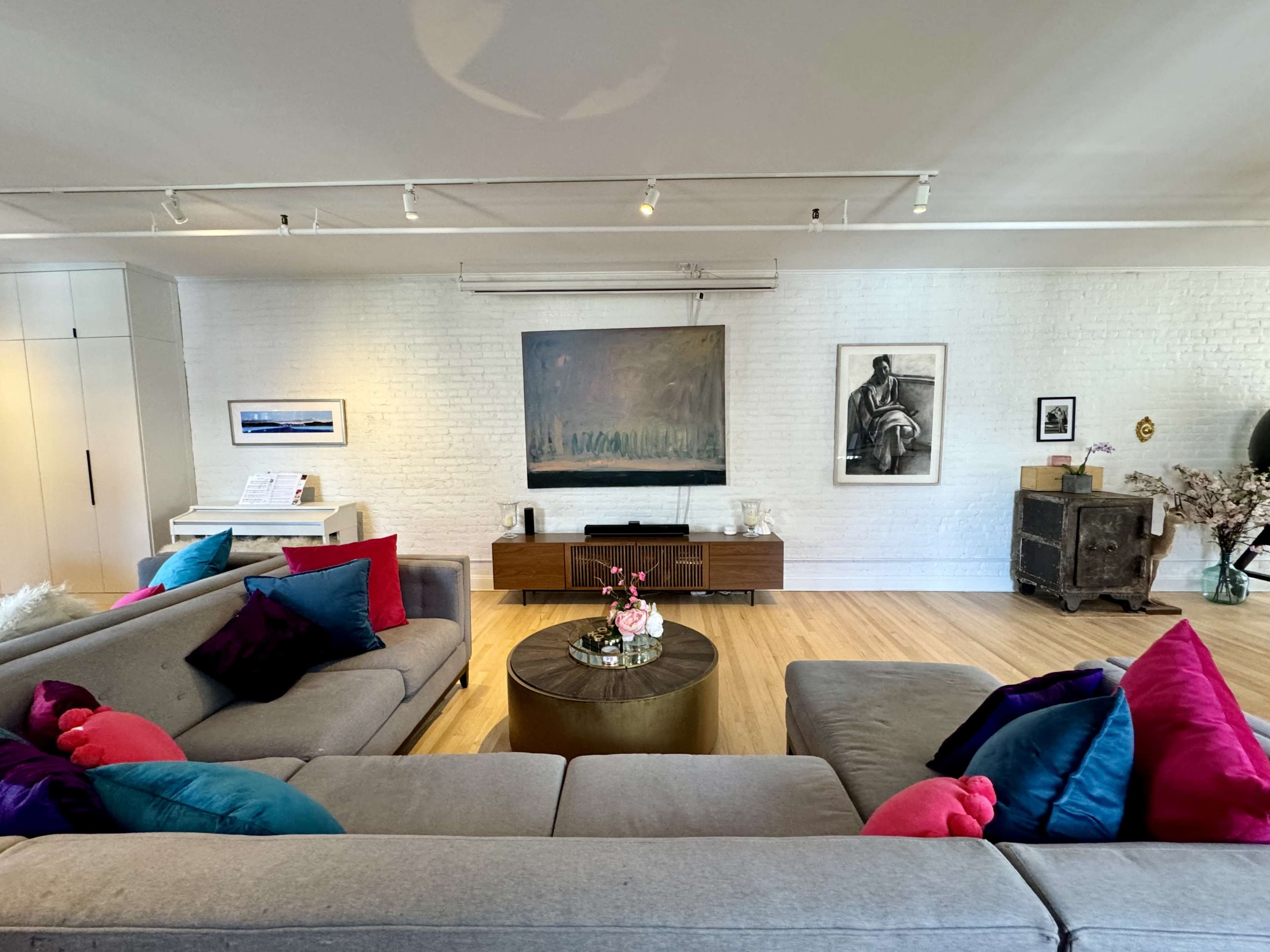 The image shows a modern living room with a gray sectional sofa adorned with colorful pillows, a round coffee table at the center, and various artworks and decorative items on the walls.