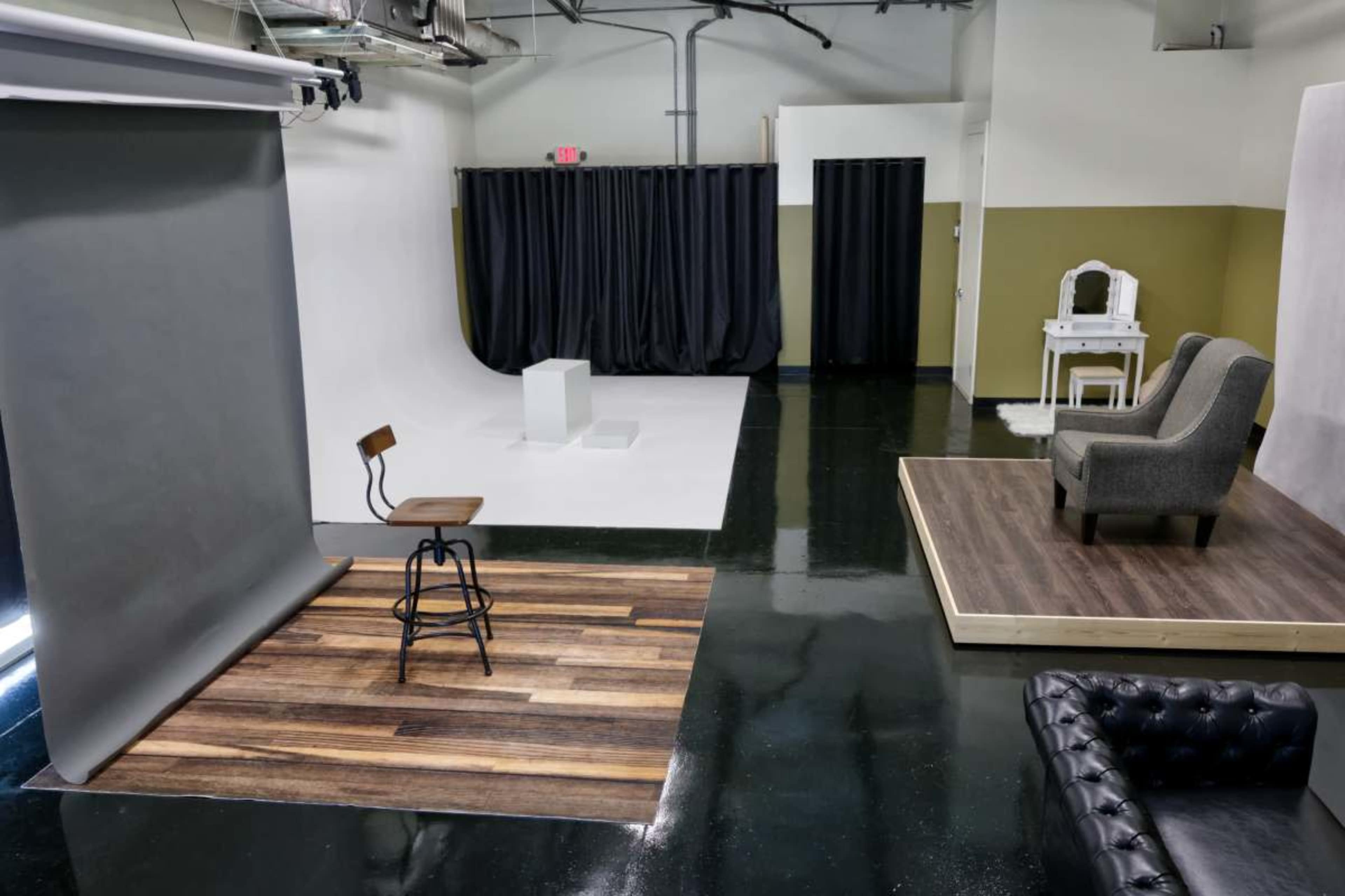 The image shows a photography studio with a gray backdrop, a wooden floor area, a black setup with curtains, and a chair positioned in front of a white space.