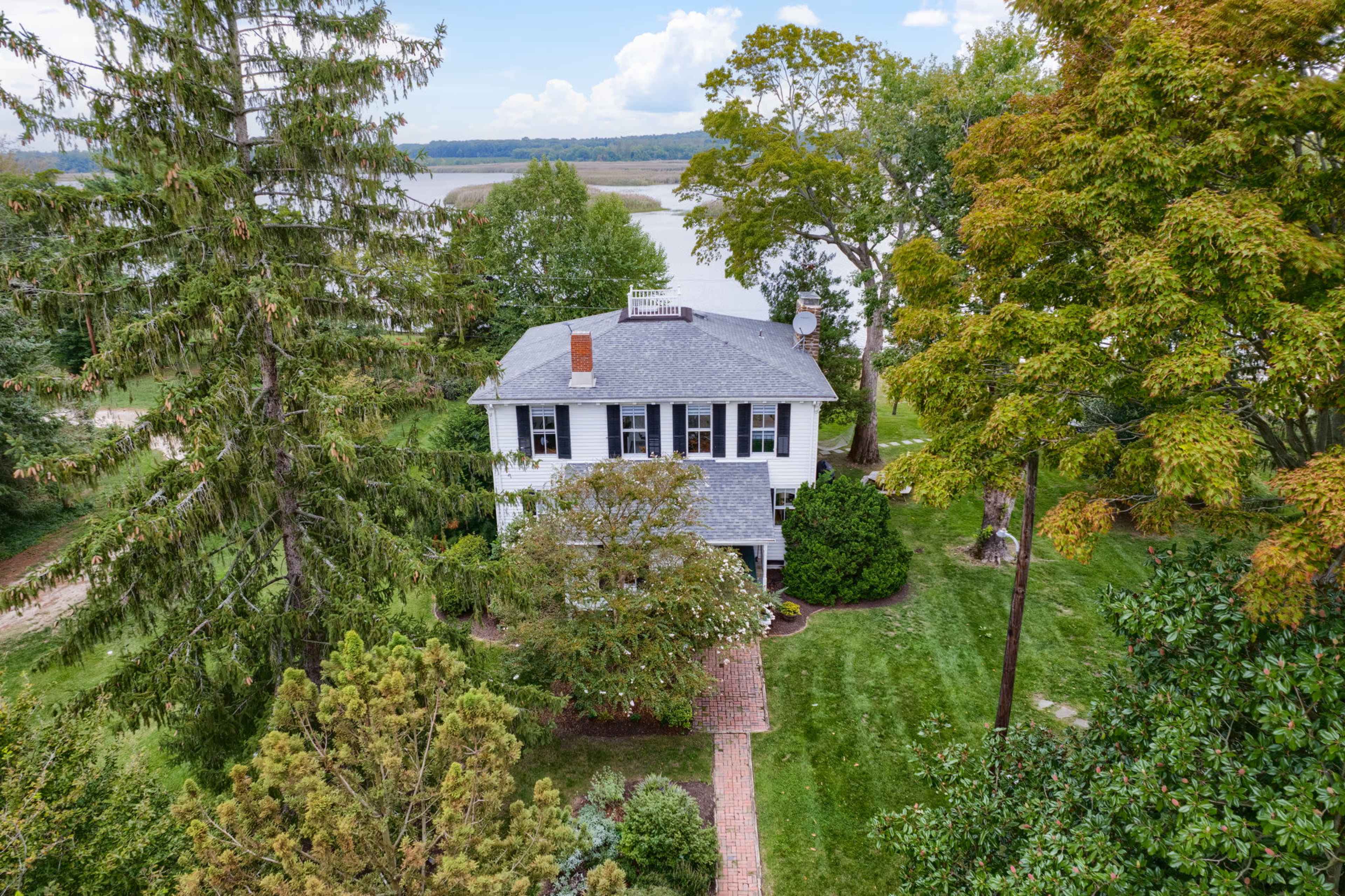 A large two-story house is surrounded by trees and a well-maintained lawn, with a river visible in the background.