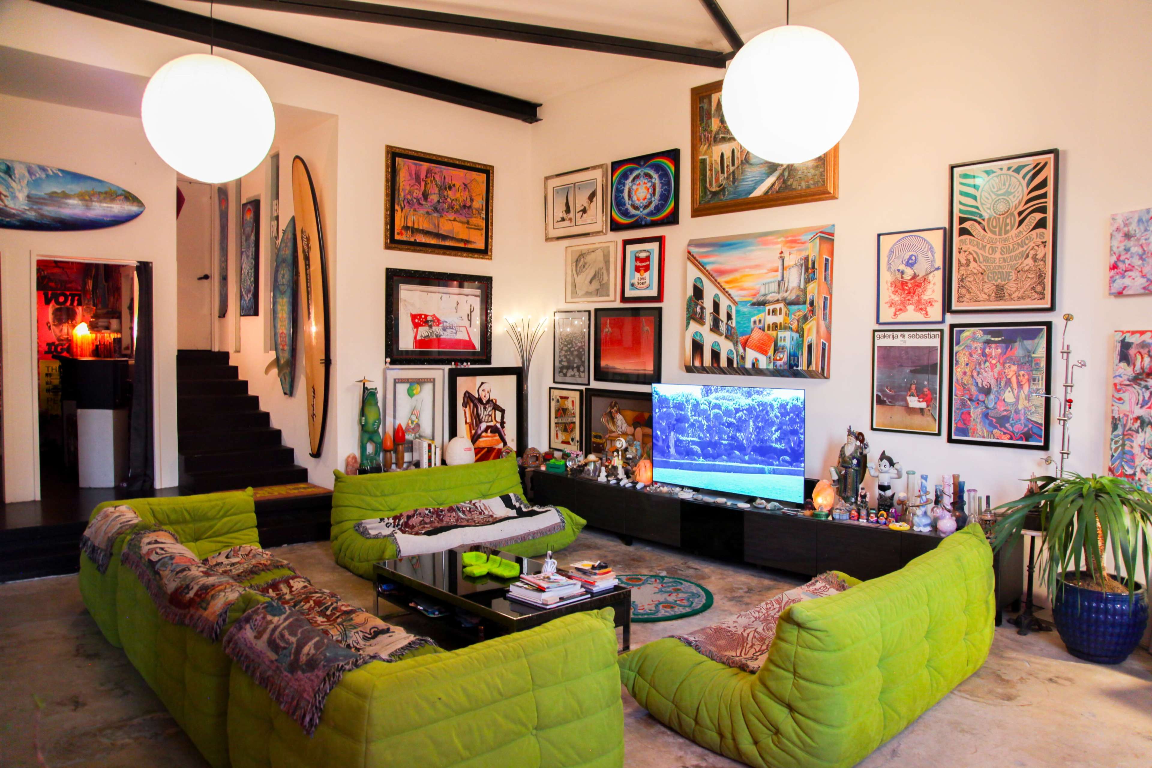 The image shows a spacious living room with a green sectional sofa, a large television, and an array of colorful artwork displayed on the walls.