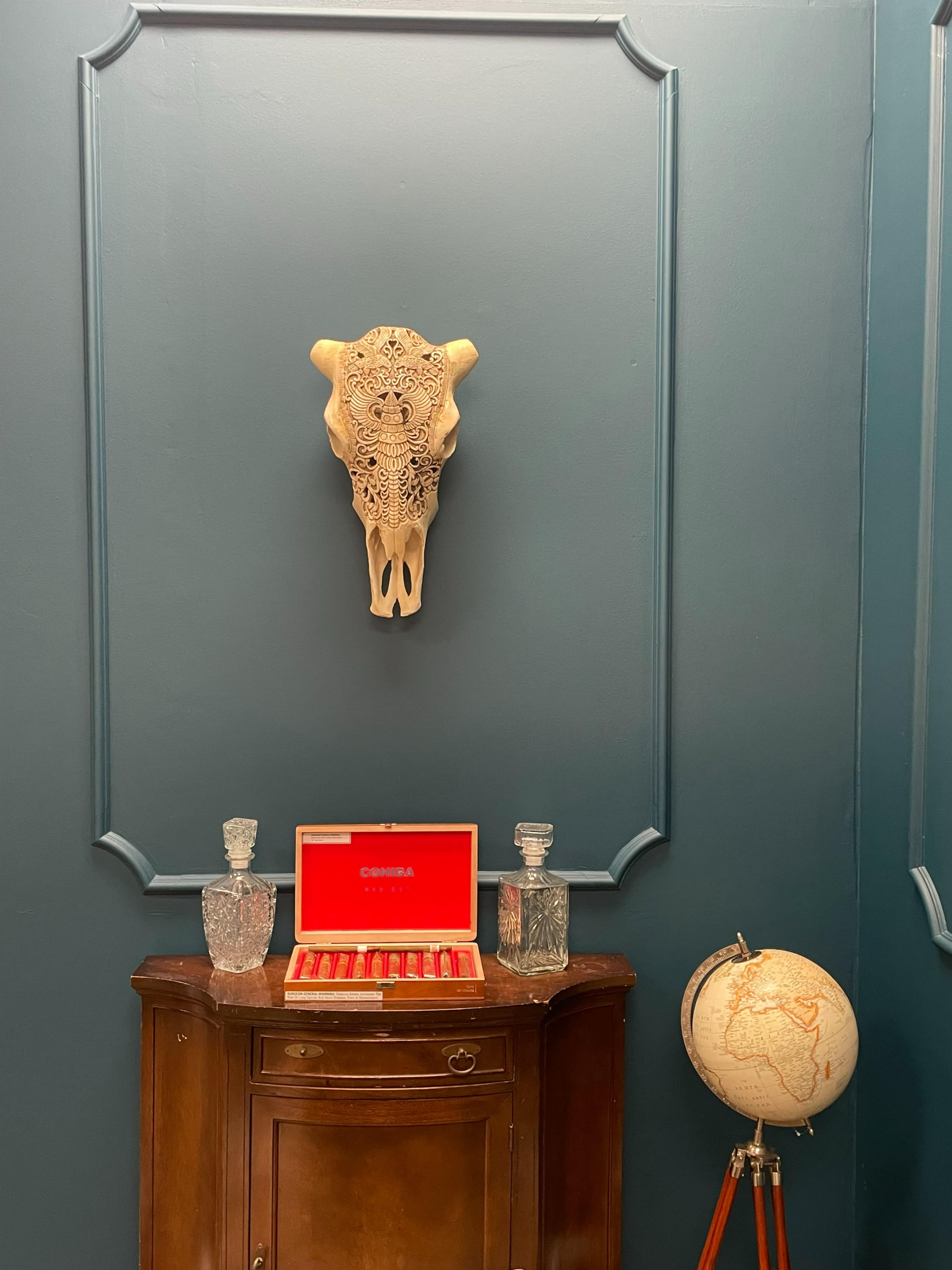 A carved skull hangs on a dark blue wall above a wooden cabinet with crystal decanters and a globe on a stand.
