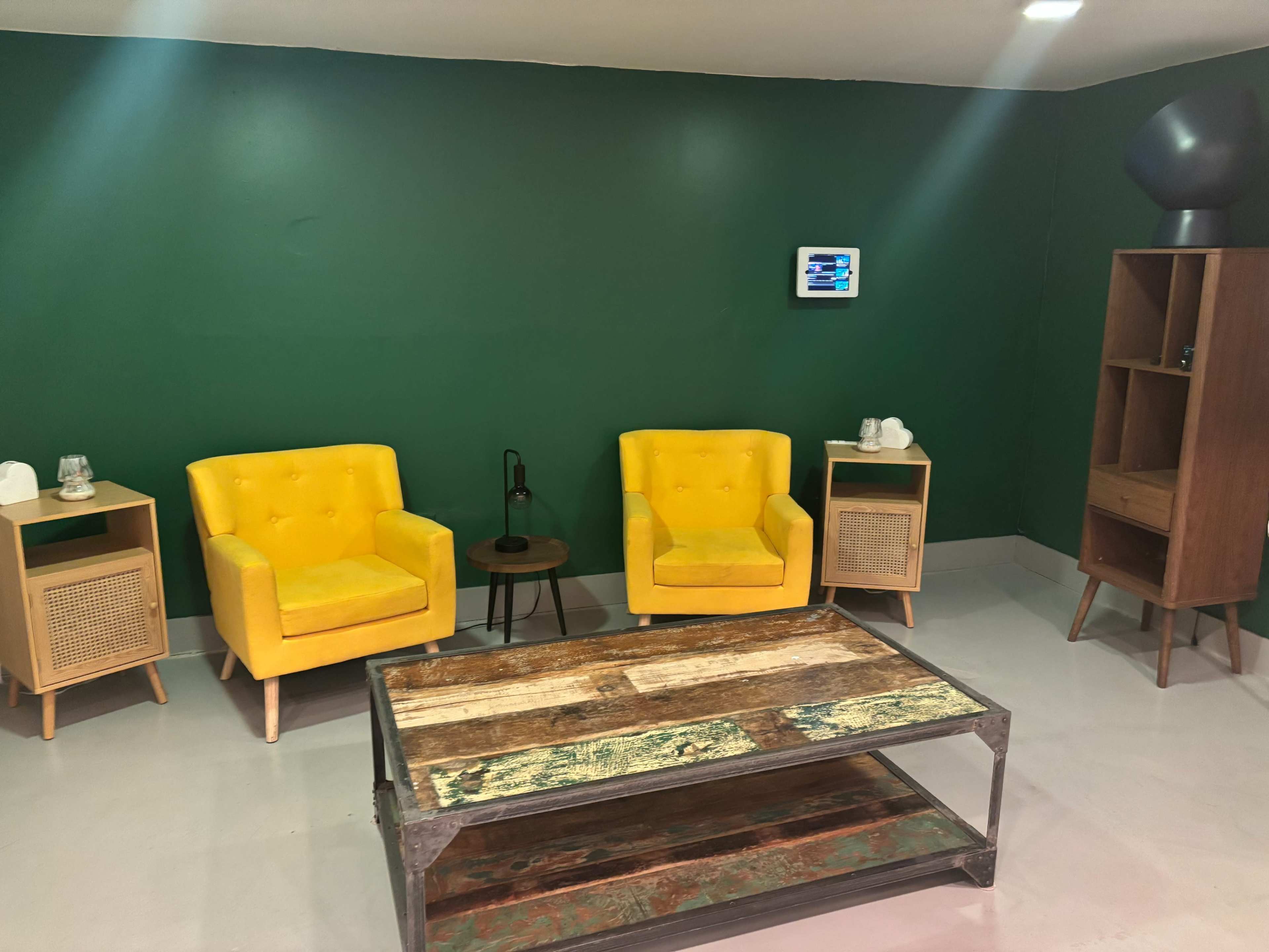 The image features a room with a green wall, two yellow armchairs, a wooden coffee table, and a cabinet in the corner.