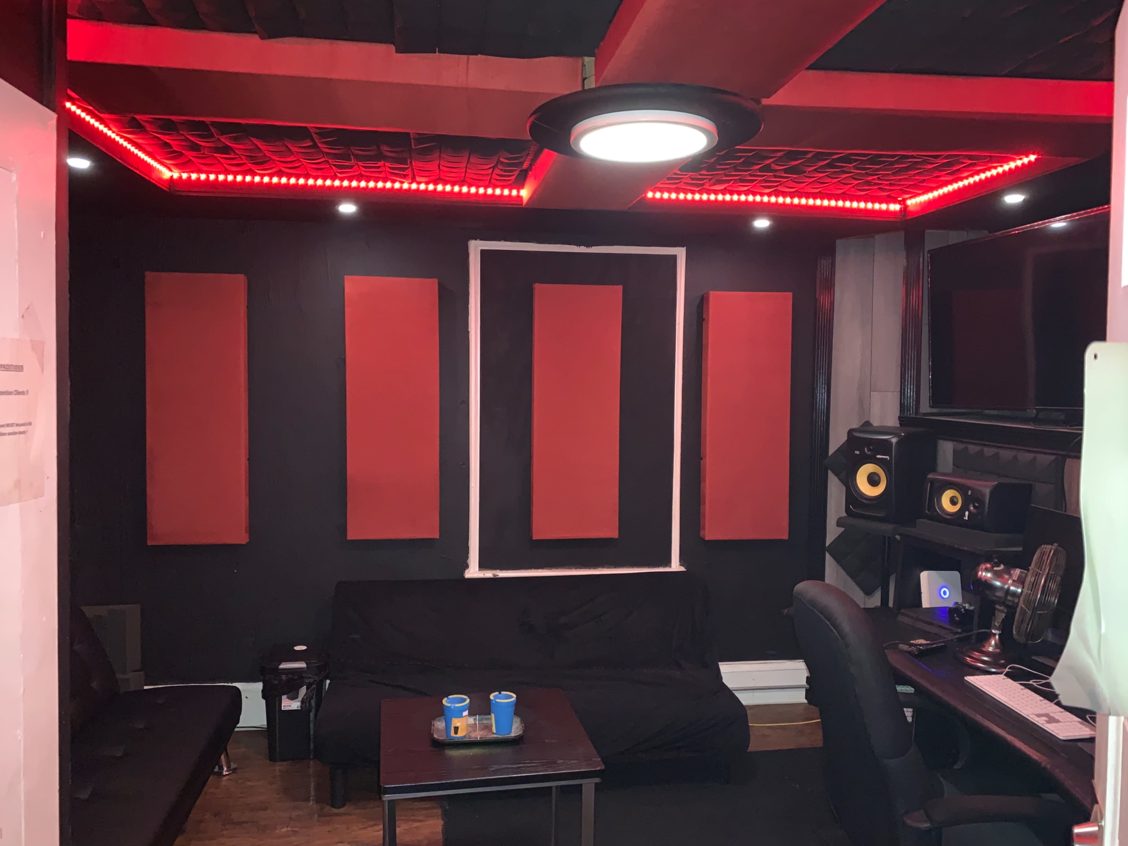 A music studio featuring black walls with red soundproofing panels, a black sofa, a small table with two cups, and audio equipment set up on a desk.