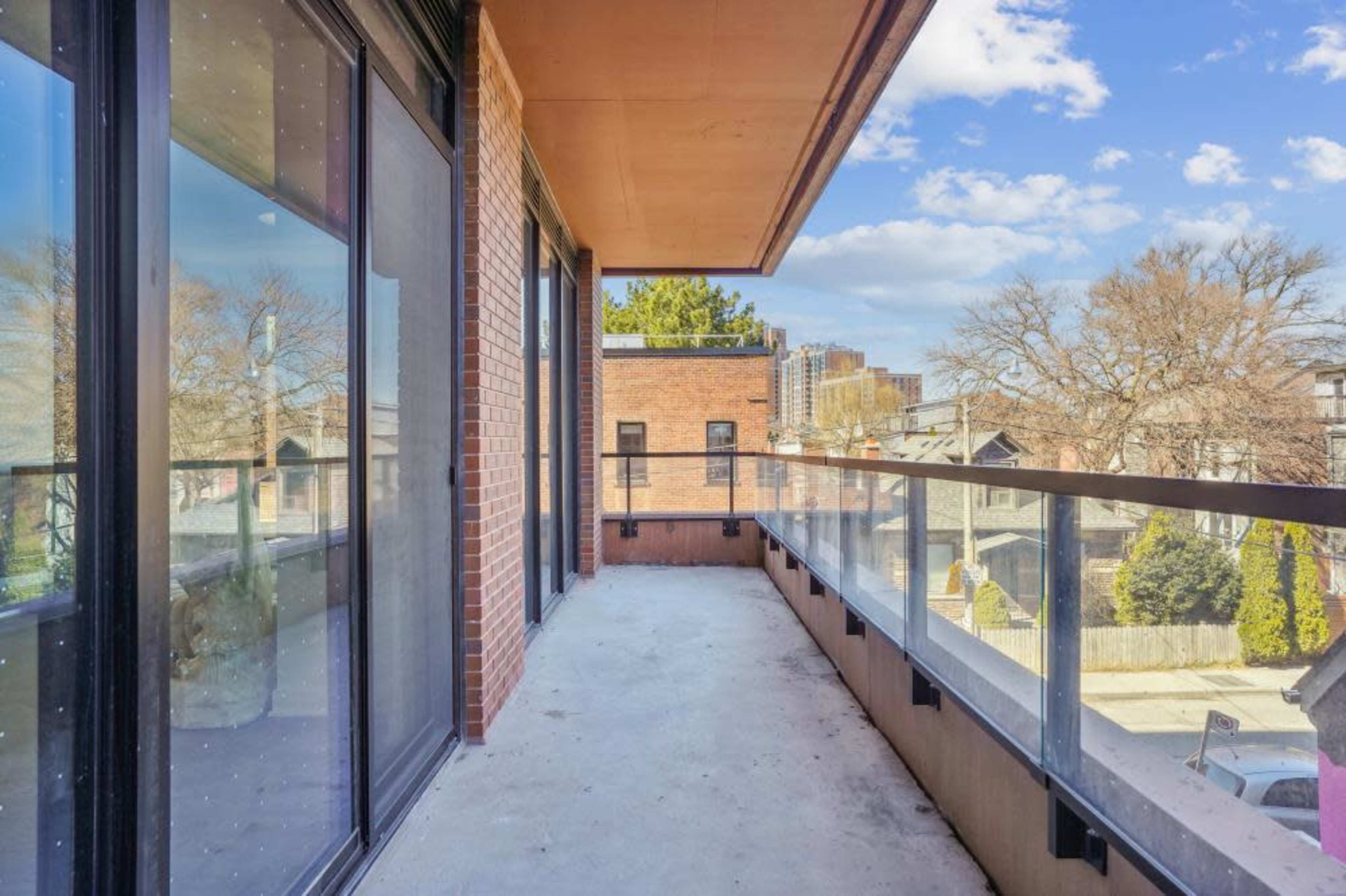 A wide balcony with glass railings overlooks a neighborhood with trees and buildings under a partly cloudy sky.